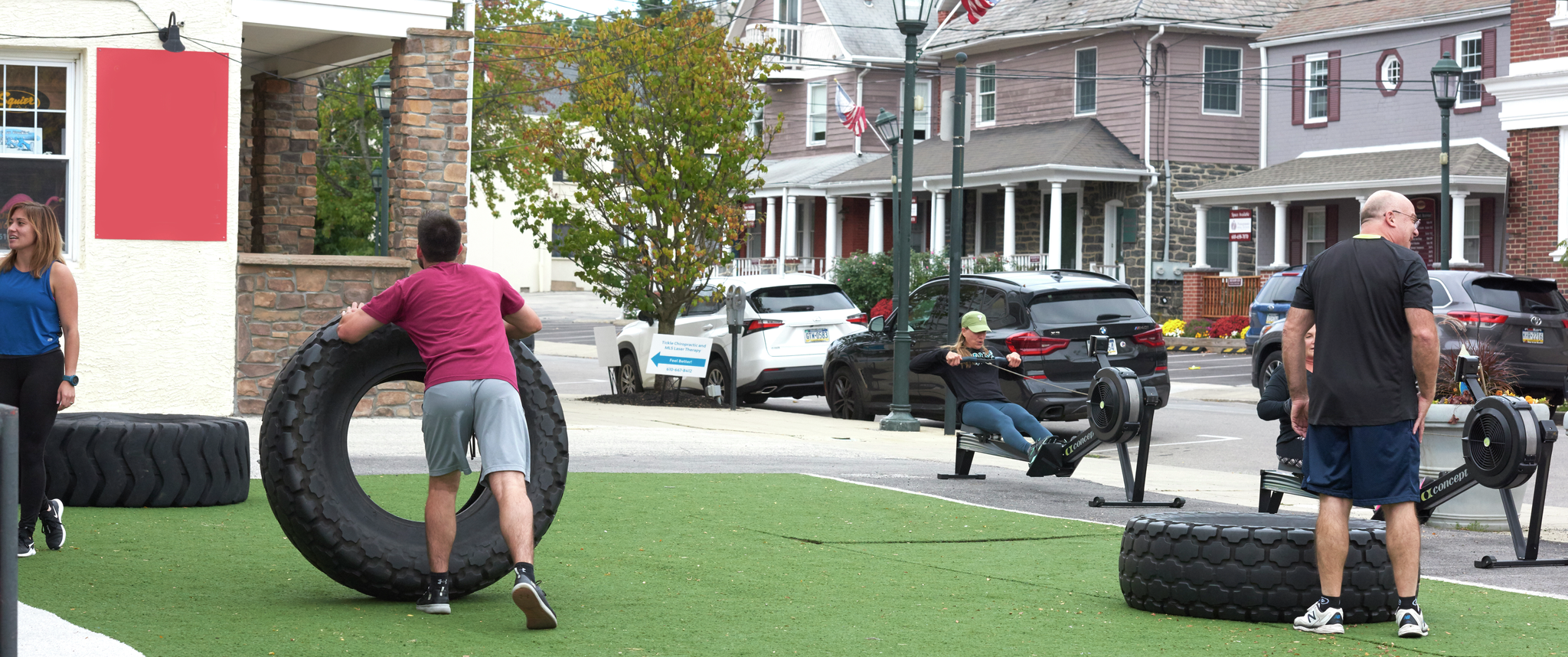 In an outdoor space in a town, several people engage in exercise with rowing machines and large tires