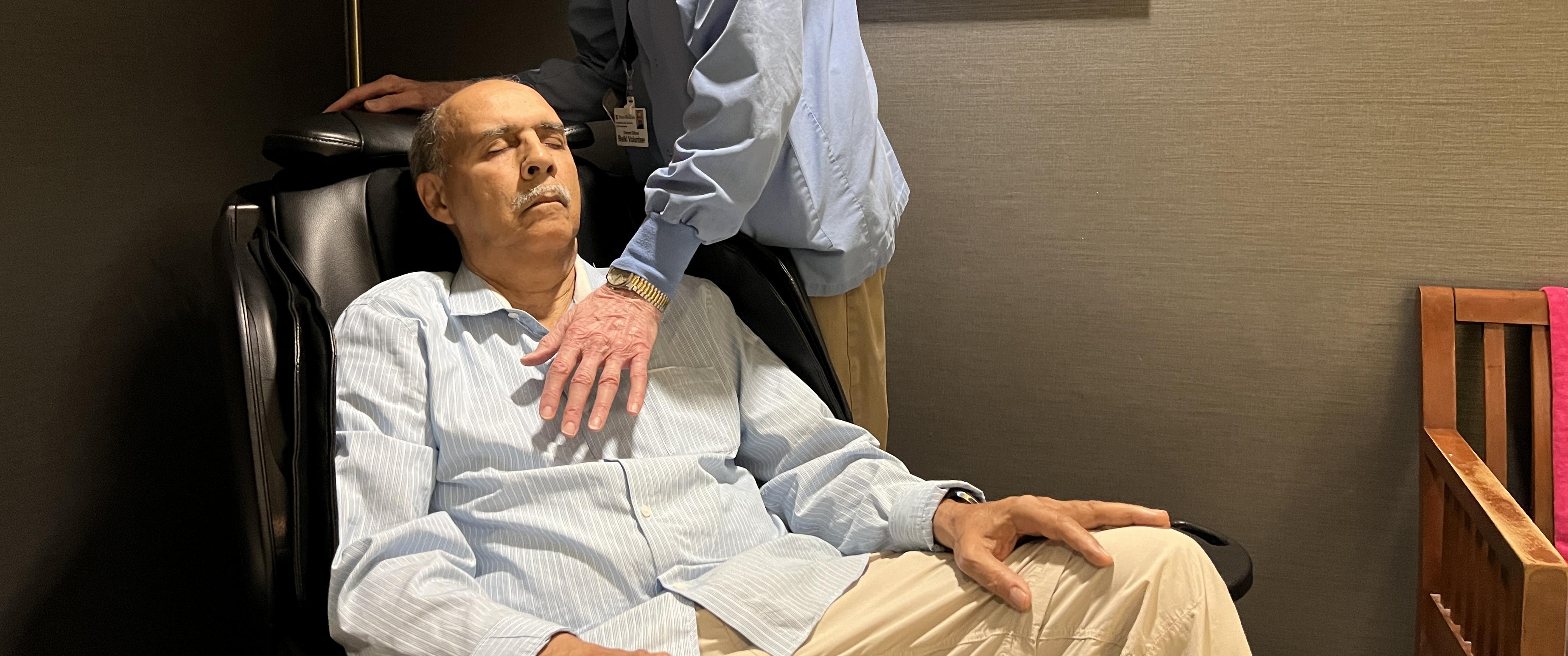 Vince Gilhool stands beside an armchair, holding his hand just above the chest of Stephen Hastings, who sits with his eyes closed