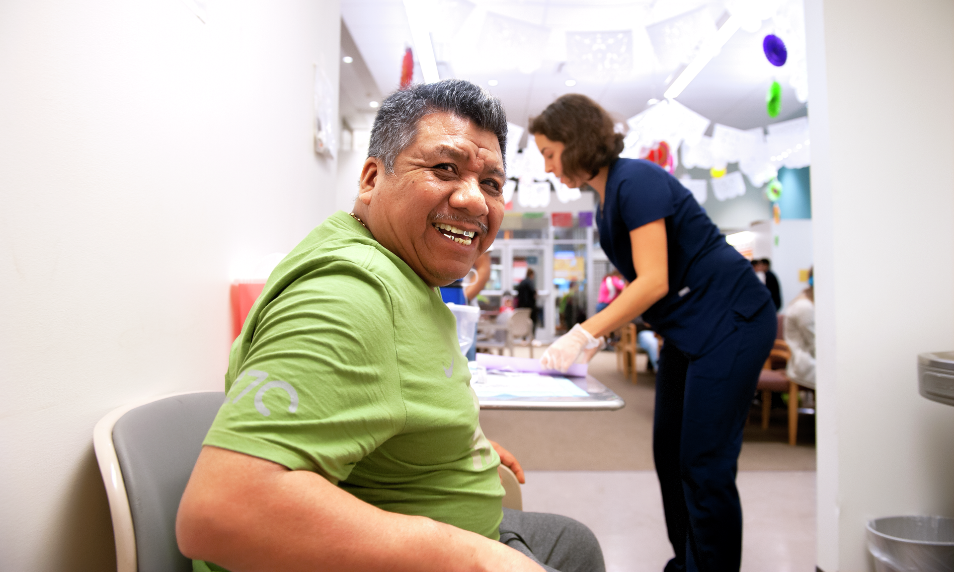 A seated middle-aged man smiles broadly while a nurse and the Puentes waiting room are in the background