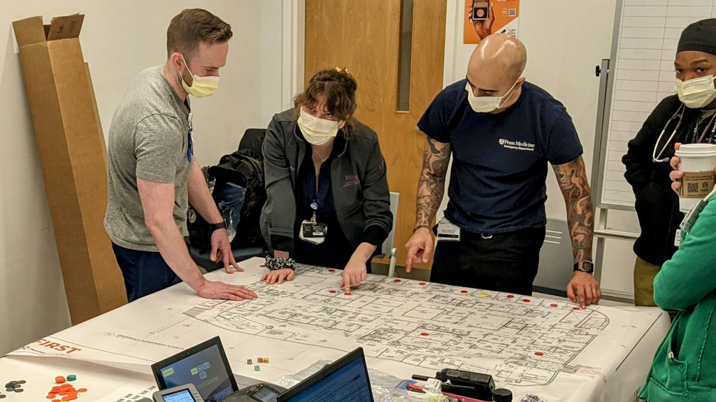 Four masked health care personnel stand around a table with a large paper map diagram spread out 
