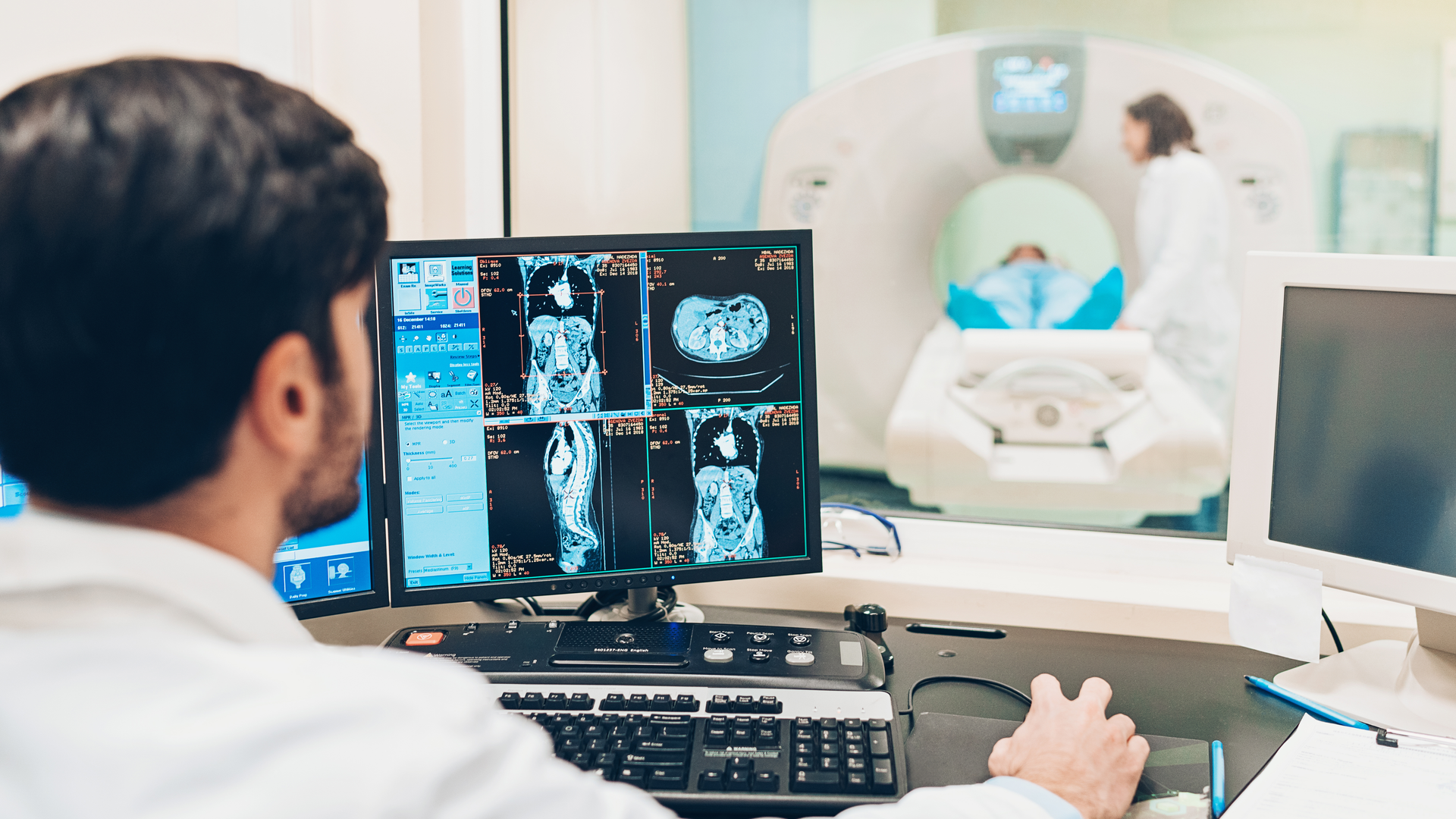 The back of a doctor looking at scans of an abdomen on his monitor while a patient recieves a CT scan in the background.