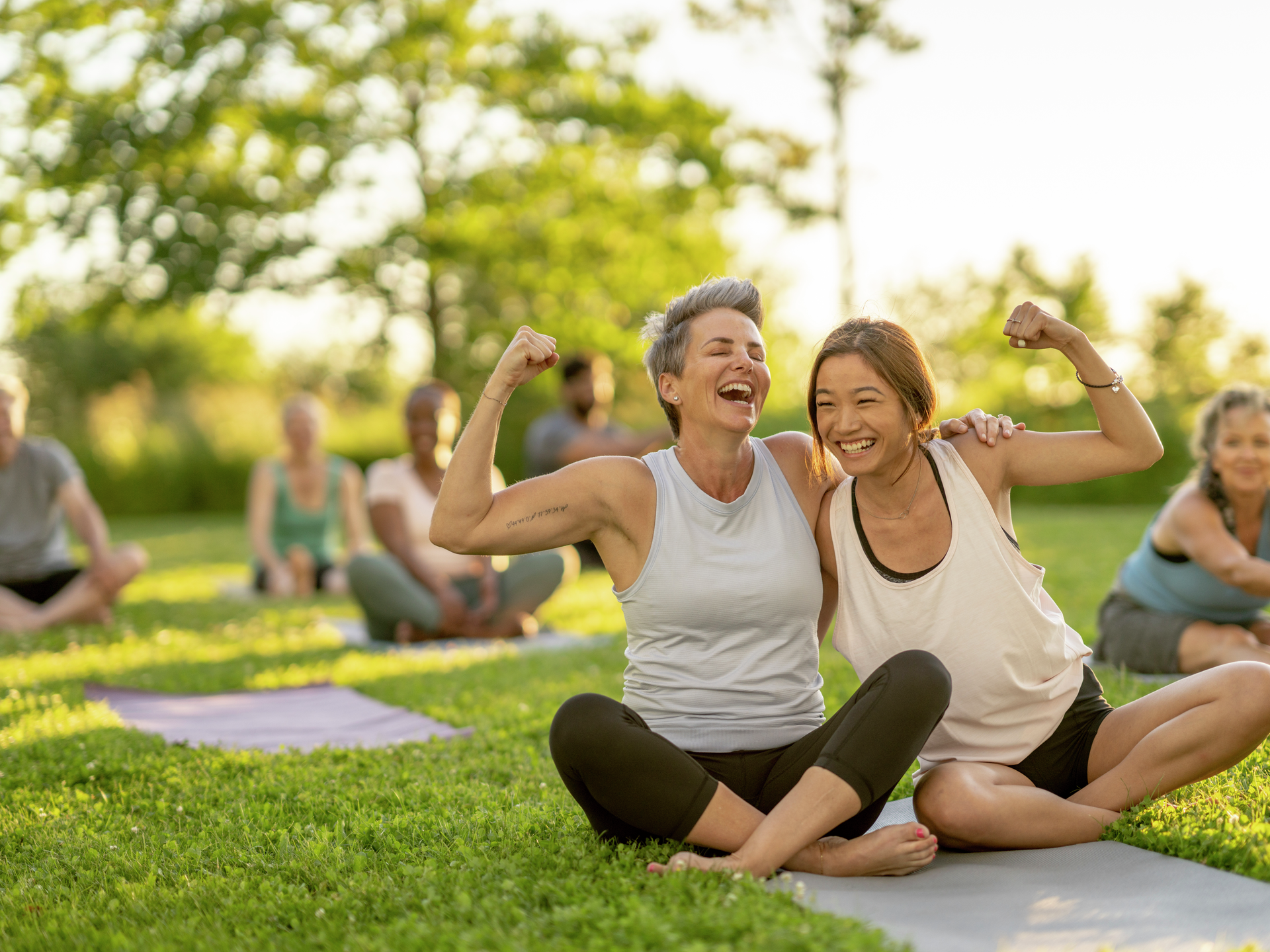 Two woman share a yoga mat as they lean in closely for a hug and flex their muscles