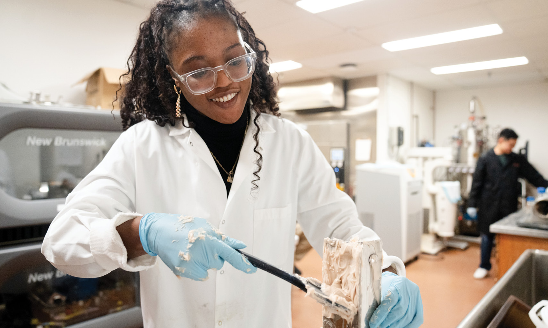 A woman in a lab coat uses a tool to scrape a mass of yeast, which resembles hummus, from a metal substrate