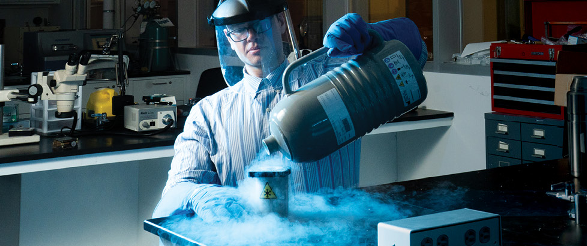 Nicholas Palmer, wearing a face shield and thick gloves, pours cold liquid nitrogen from a jug into a vessel with a snowflake symbol in a coolly lit lab