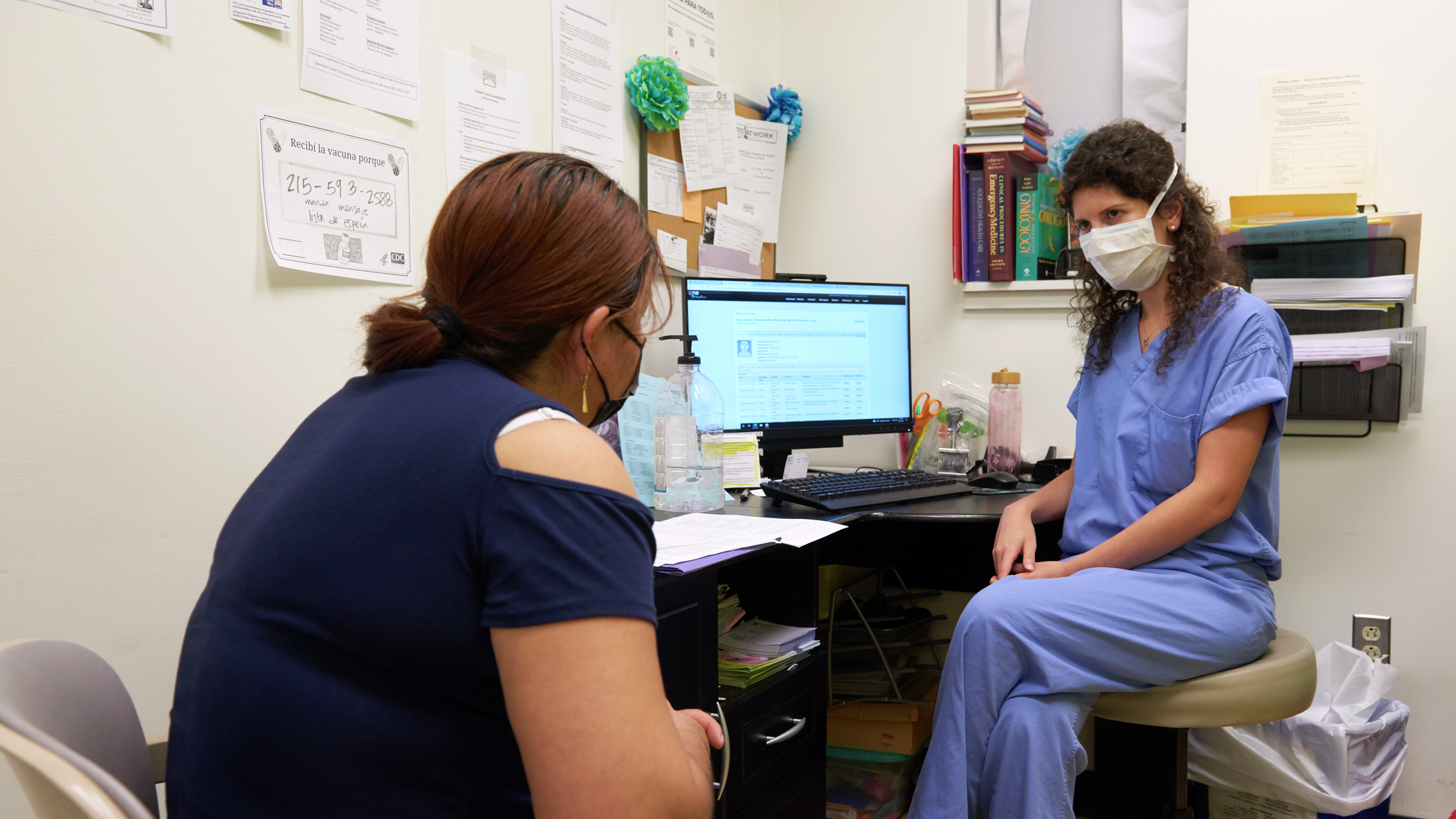 A medical student in a surgical mask meets with a middle-aged female patient in an exam room