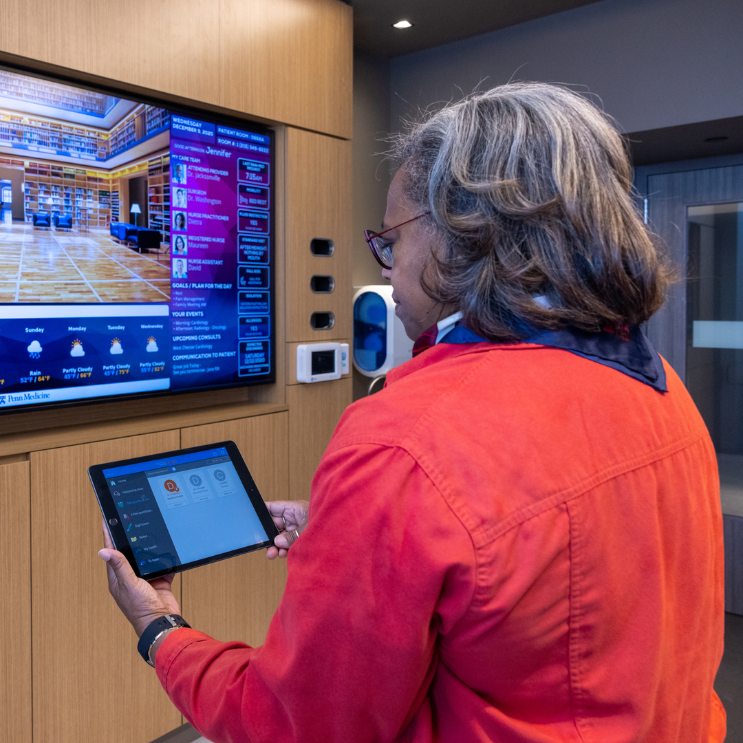 Poppy Bass holds a tablet and faces a large-screen TV in a hospital room
