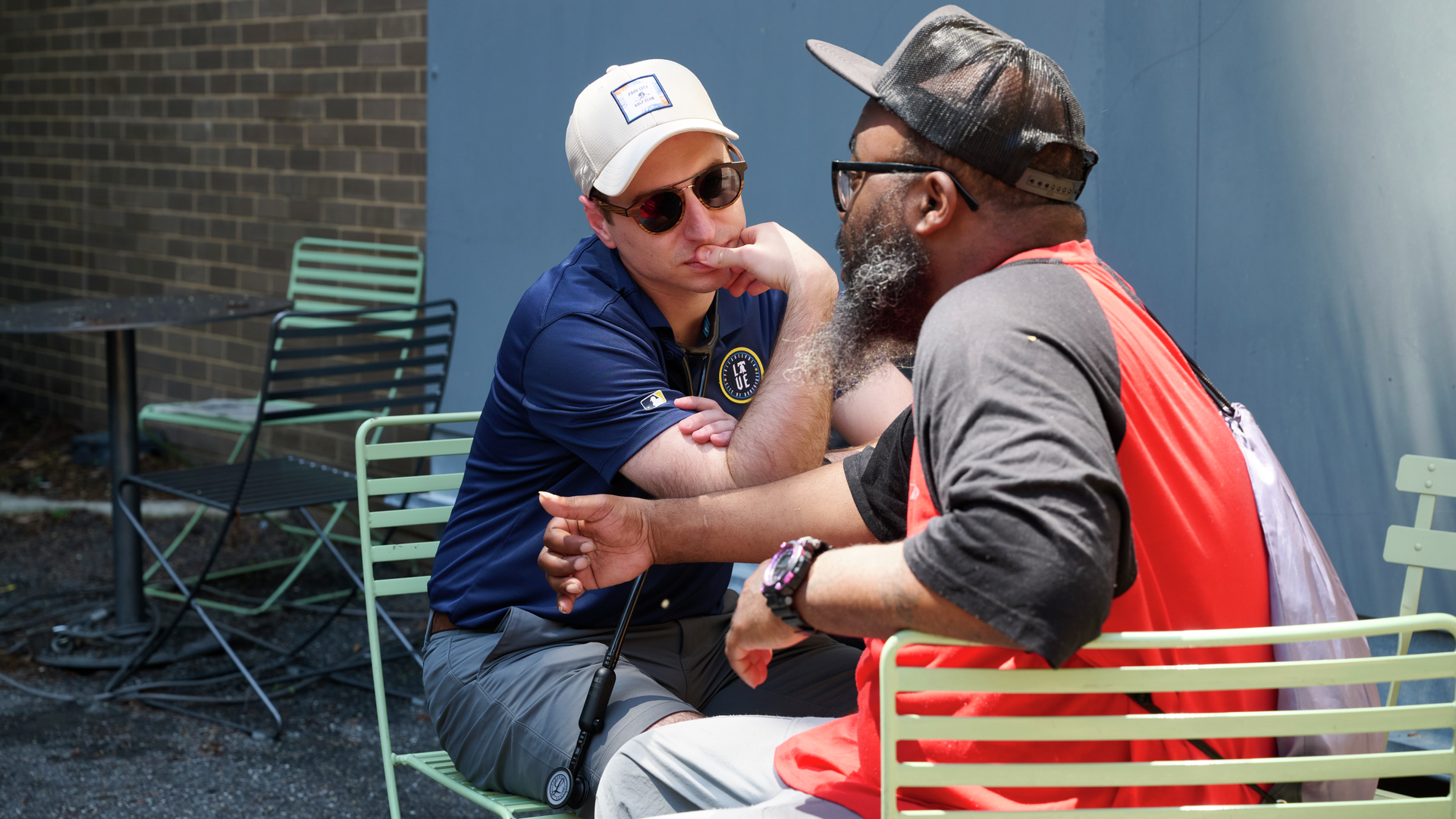 At a courtyard table, family physician Zachary Zosak, MD, sits with a bearded man wearing a hat and an orange and black shirt