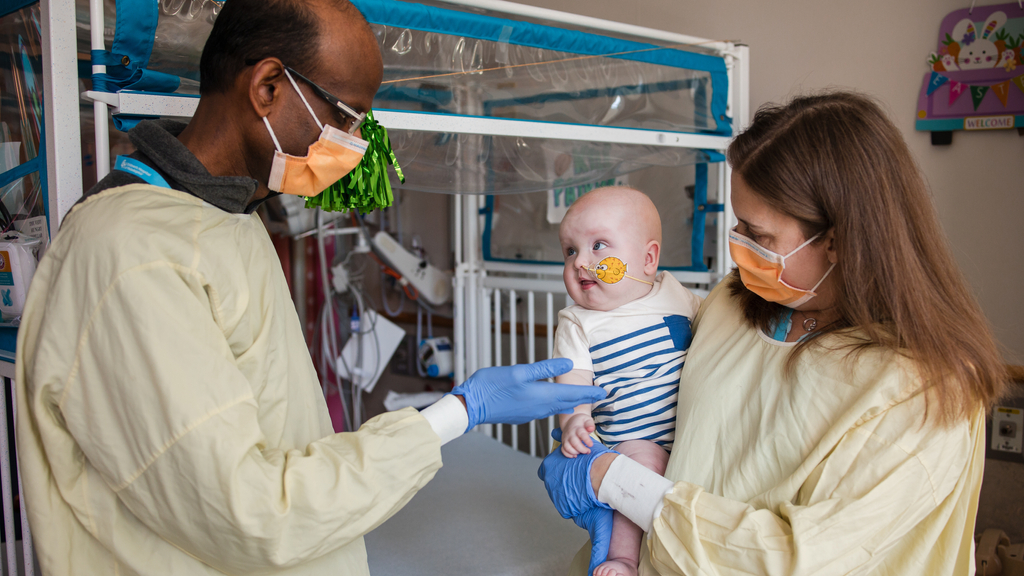 Rebecca Ahrens-Nicklas holds baby KJ while Kiran Musunuru gestures toward him; both adults masked and gowned