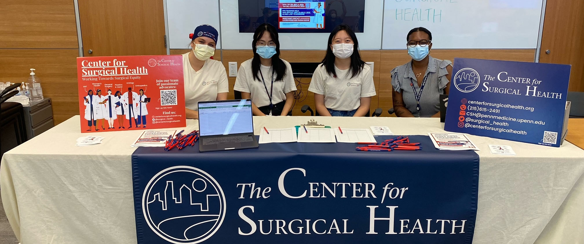 Three volunteers sit behind a desk with a Center for Surgical Health banner