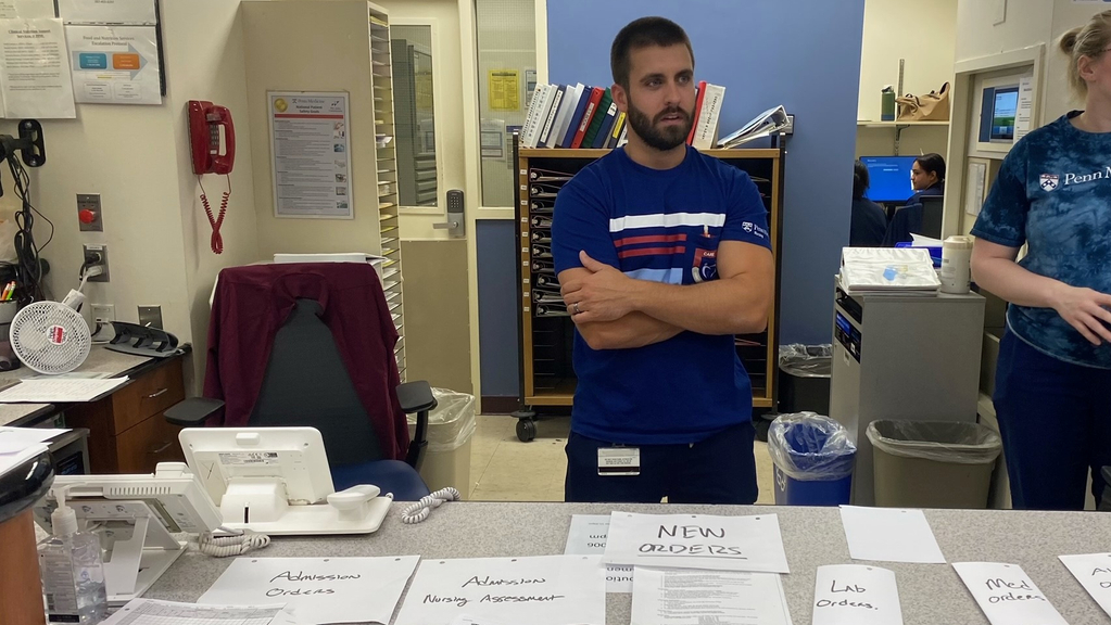 Mike Tomasetti, a nurse manager at Penn Presbyterian Medical Center, stands at a nurse’s station covered with stacks of paper orders