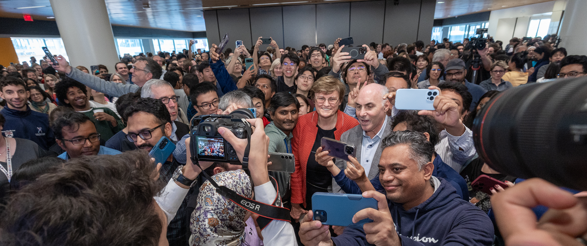 A massive crowd of people stands tightly packed around Katalin Kariko and Drew Weissman at the center, with many people taking photos of or selfies with them