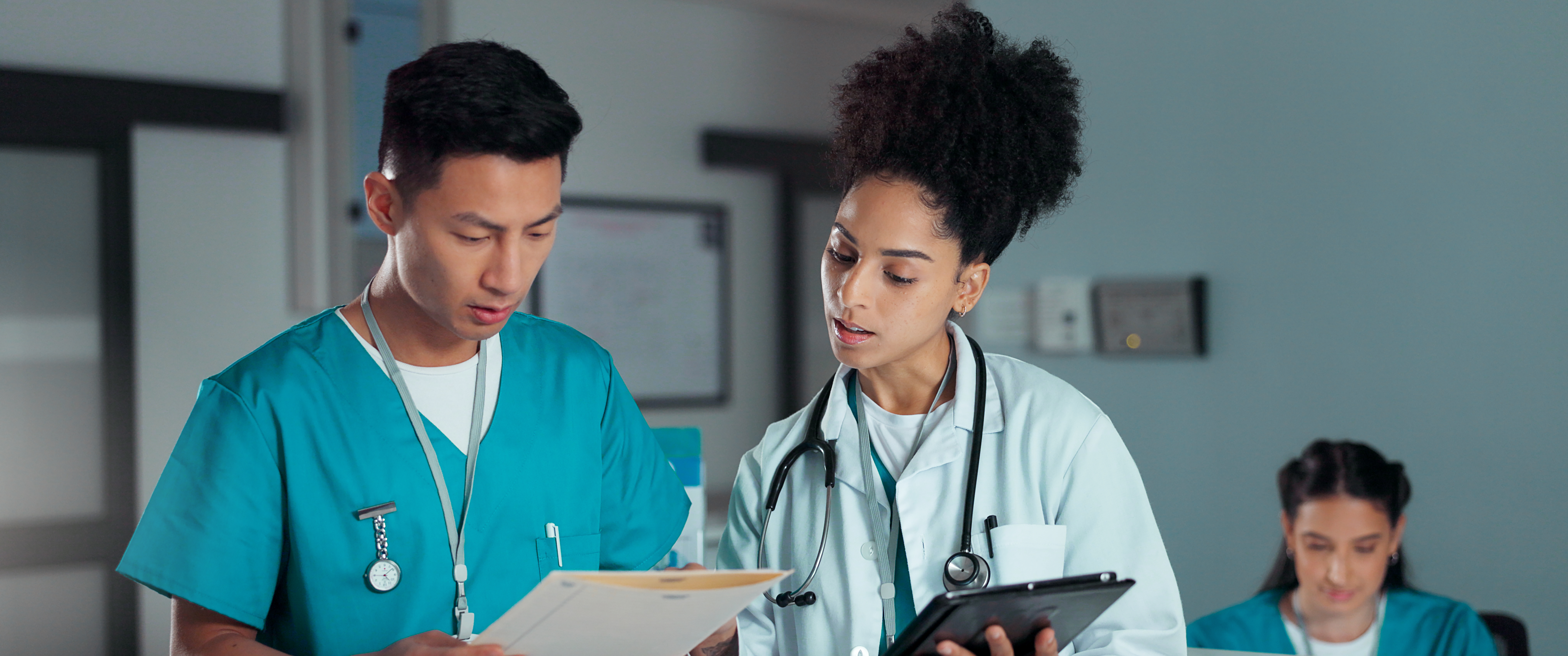 A doctor and a nurse stand together looking at a paper chart and a tablet, seeming slightly confused