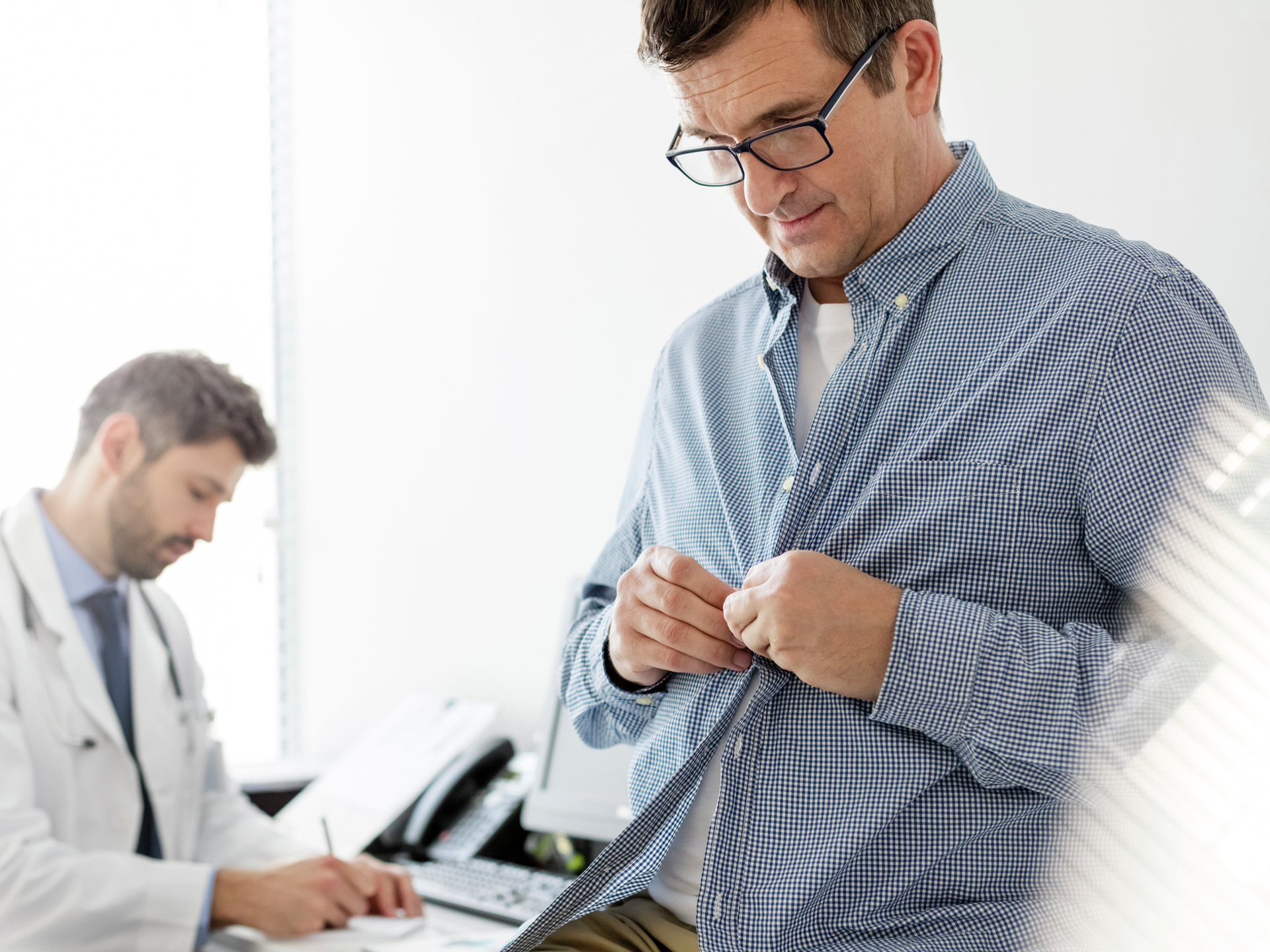 Patient buttons up his shirt after a checkup while doctor is writing a medical report at a hospital.
