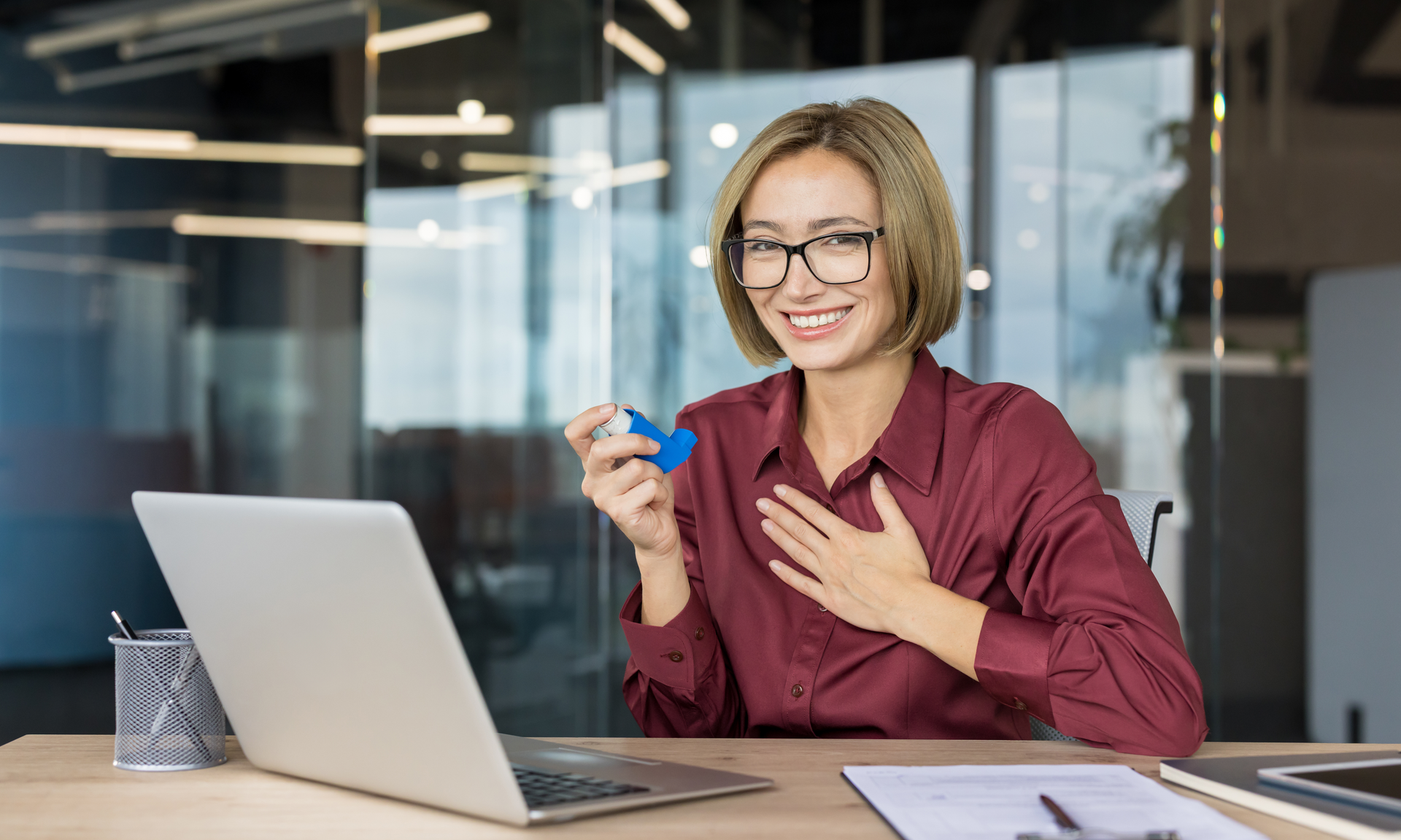 Young businesswoman smiling, feeling relief after using a blue asthma inhaler, managing her chronic obstructive pulmonary disease in her modern office workplace.