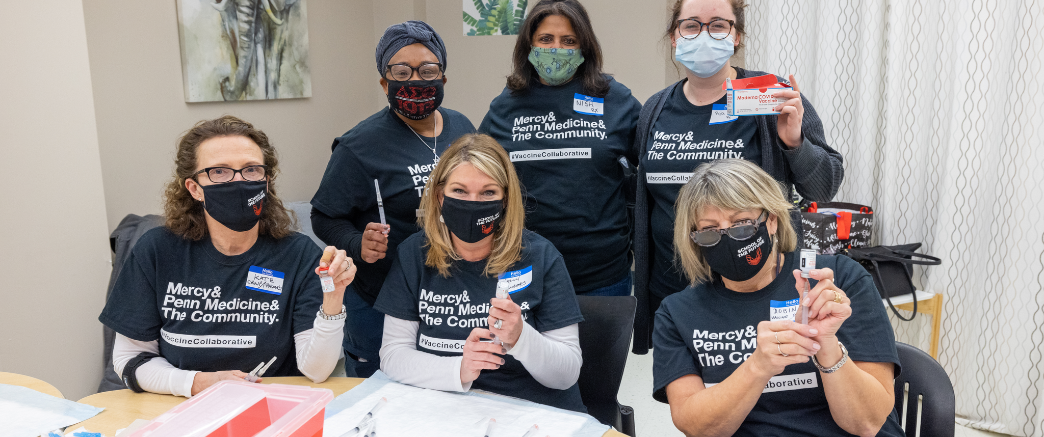A group of clinicians wearing matching T-shirts appear to be smiling beneath their masks, as they hold up vials and syringes