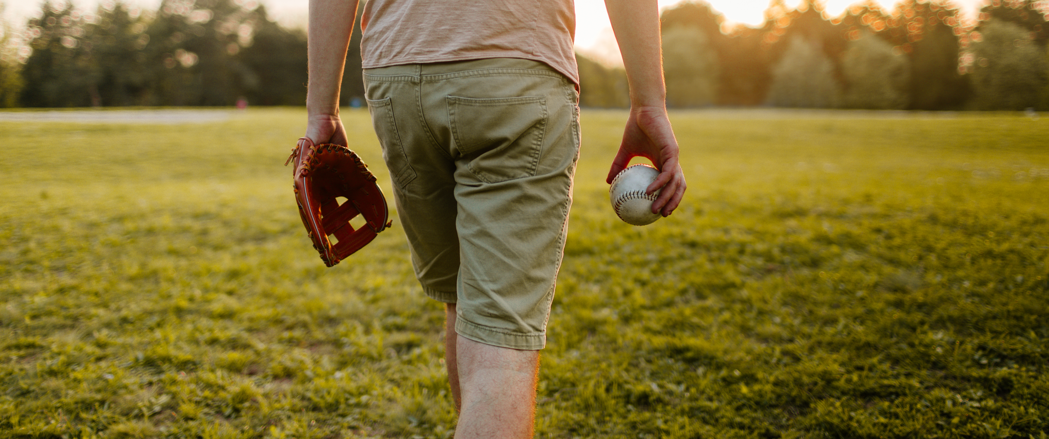 Man holding a baseball and baseball glove, walking on a field at sunset