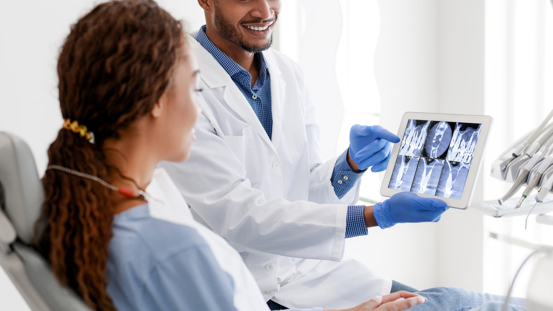 A dentist smiling while showing his patient x-rays of her teeth on a tablet.