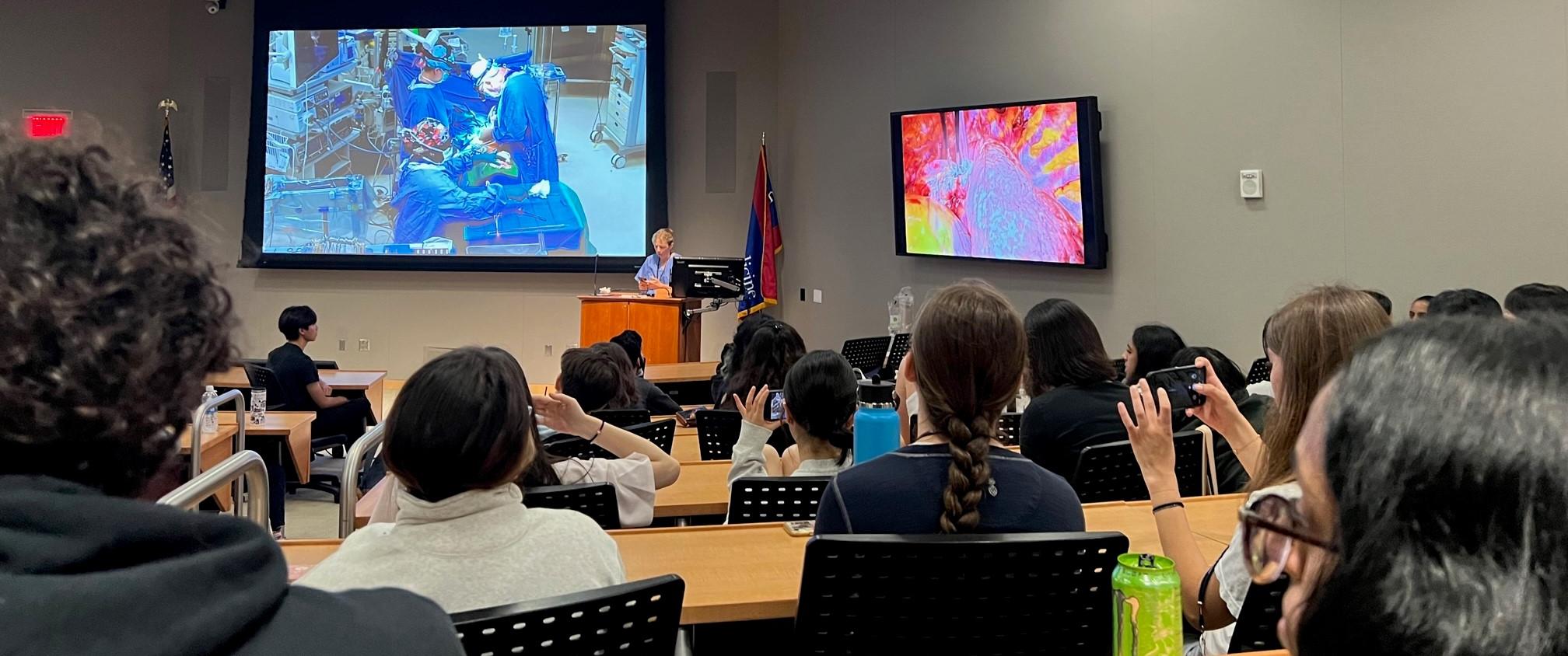 A group of students seen from behind in a classroom watch a surgical procedure on a large TV screen