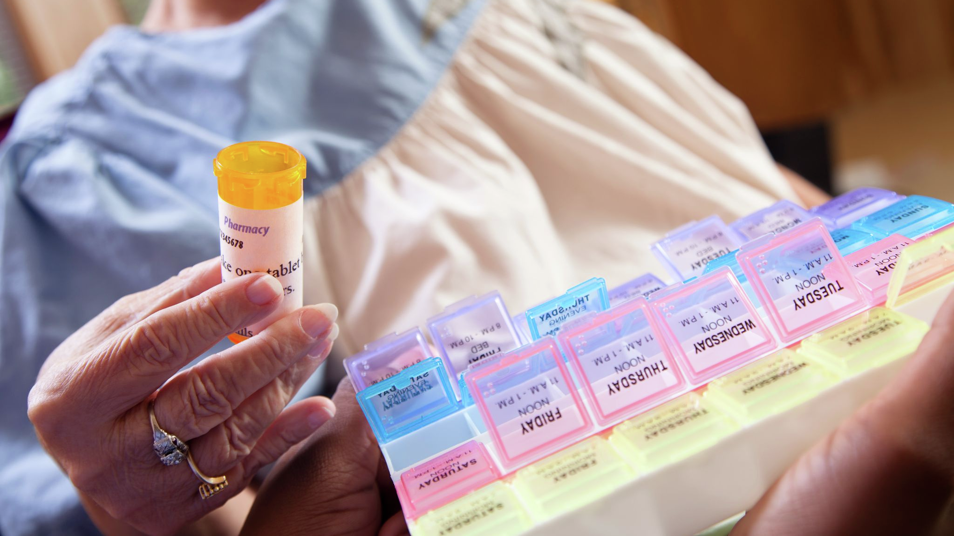 A senior woman wearing a hospital gown holds an orange pill bottle in front of a daily pill case container.