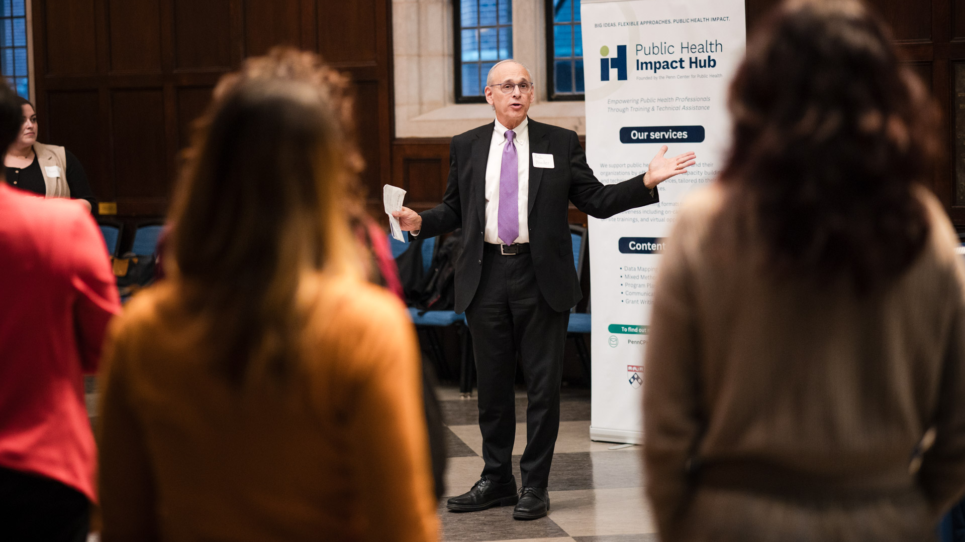Richard Wender speaking in front of a group of people with a poster announcing the Impact Hub in the background