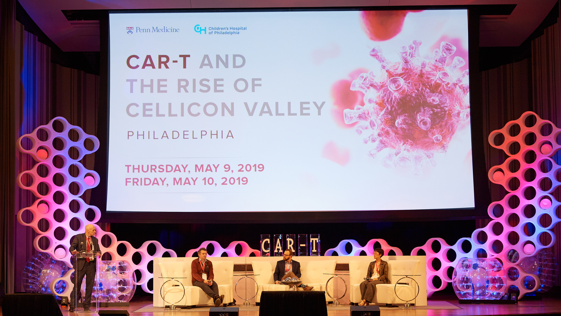 Carl June stands at a podium on a stage with modern, cell-themed furnishings, with three seated panelists and a large screen that says "Cellicon Valley"