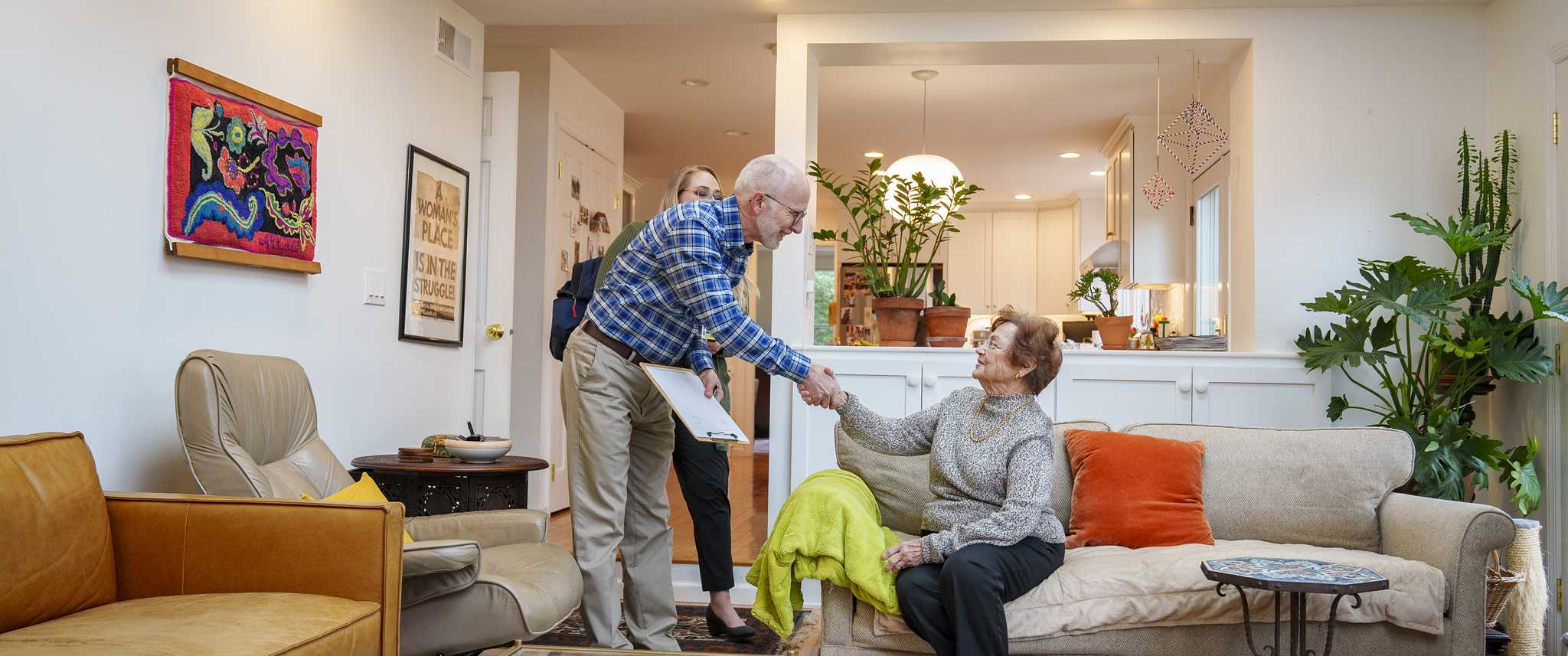 Leon Kraybill, a smiling man with white beard, stands and shakes the hand of an older woman seated on a comfortable home couch.