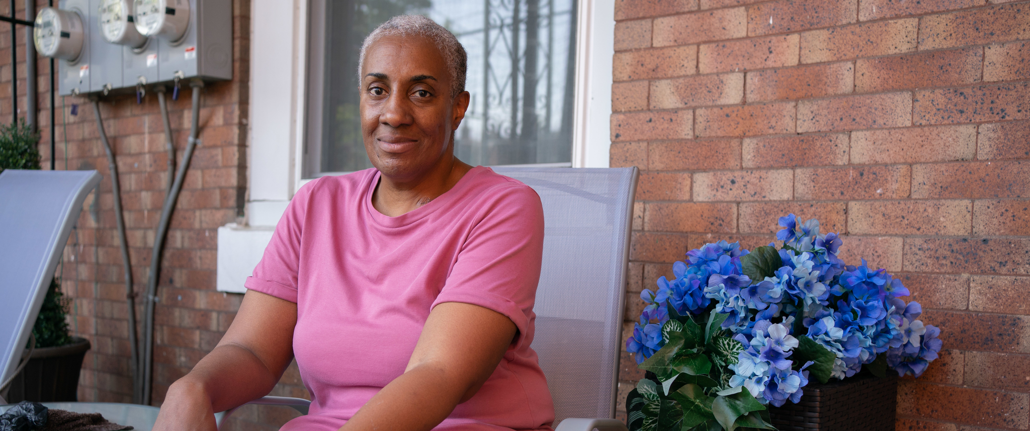 Rubella Utendhal, a woman with close-cropped gray hair wearing a pink dress, sits on her front porch