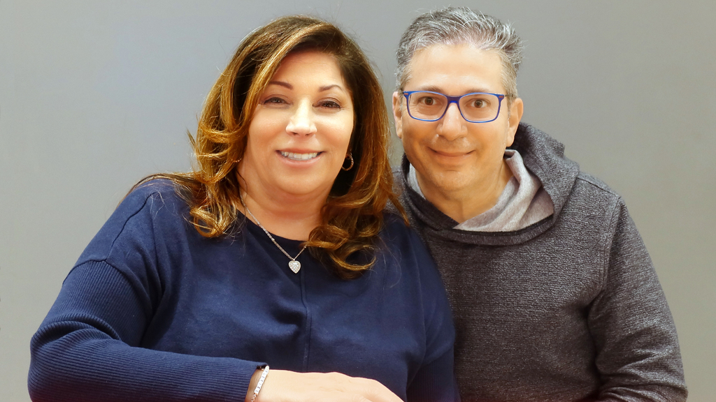 Middle-aged woman with long brown hair wearing blue sweater sitting beside man with glasses wearing grey sweatshirt.