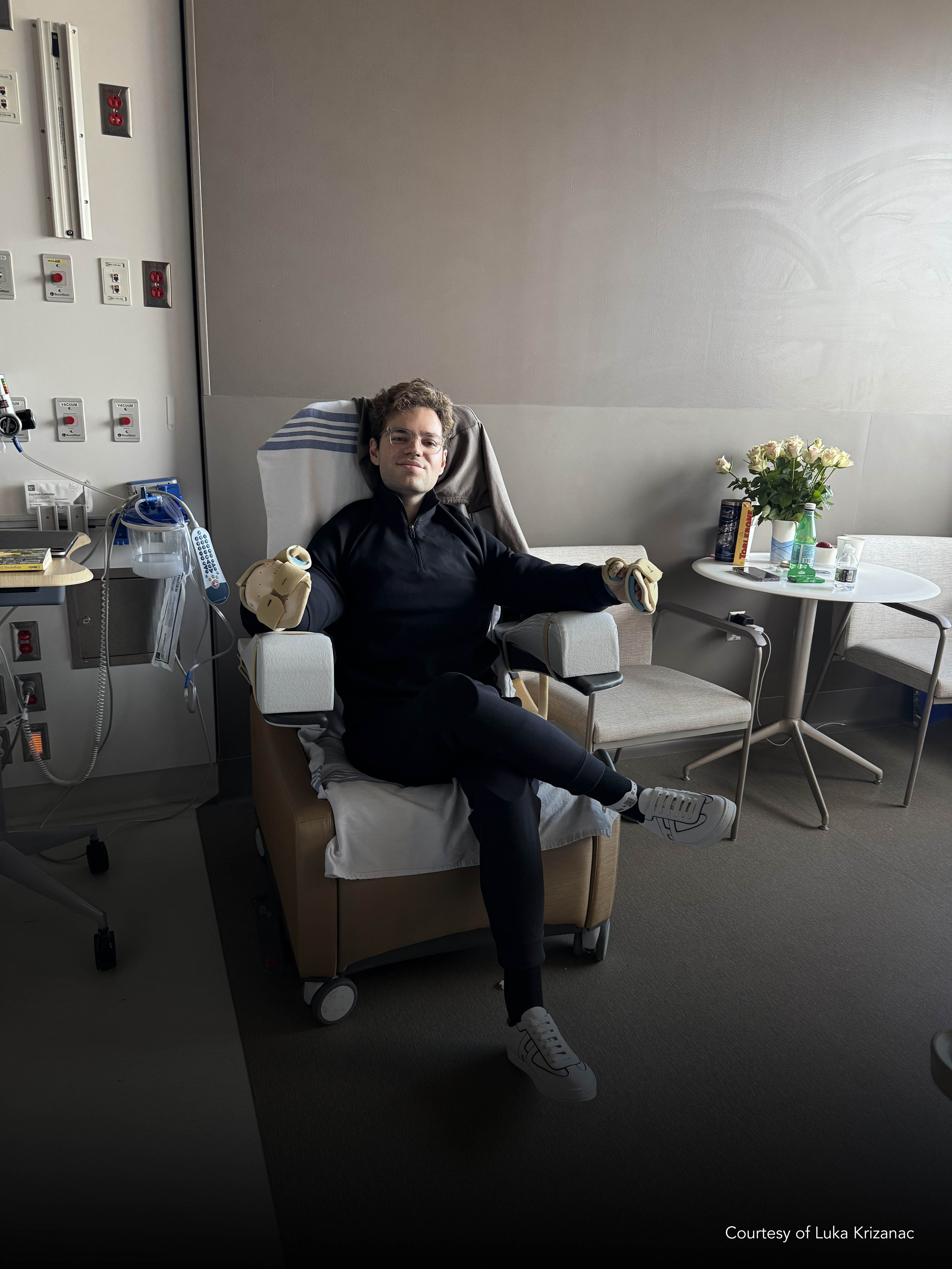 Luka Krizanac sitting in a chair in his patient room with casts on his hands and text of a photo courtesy in corner