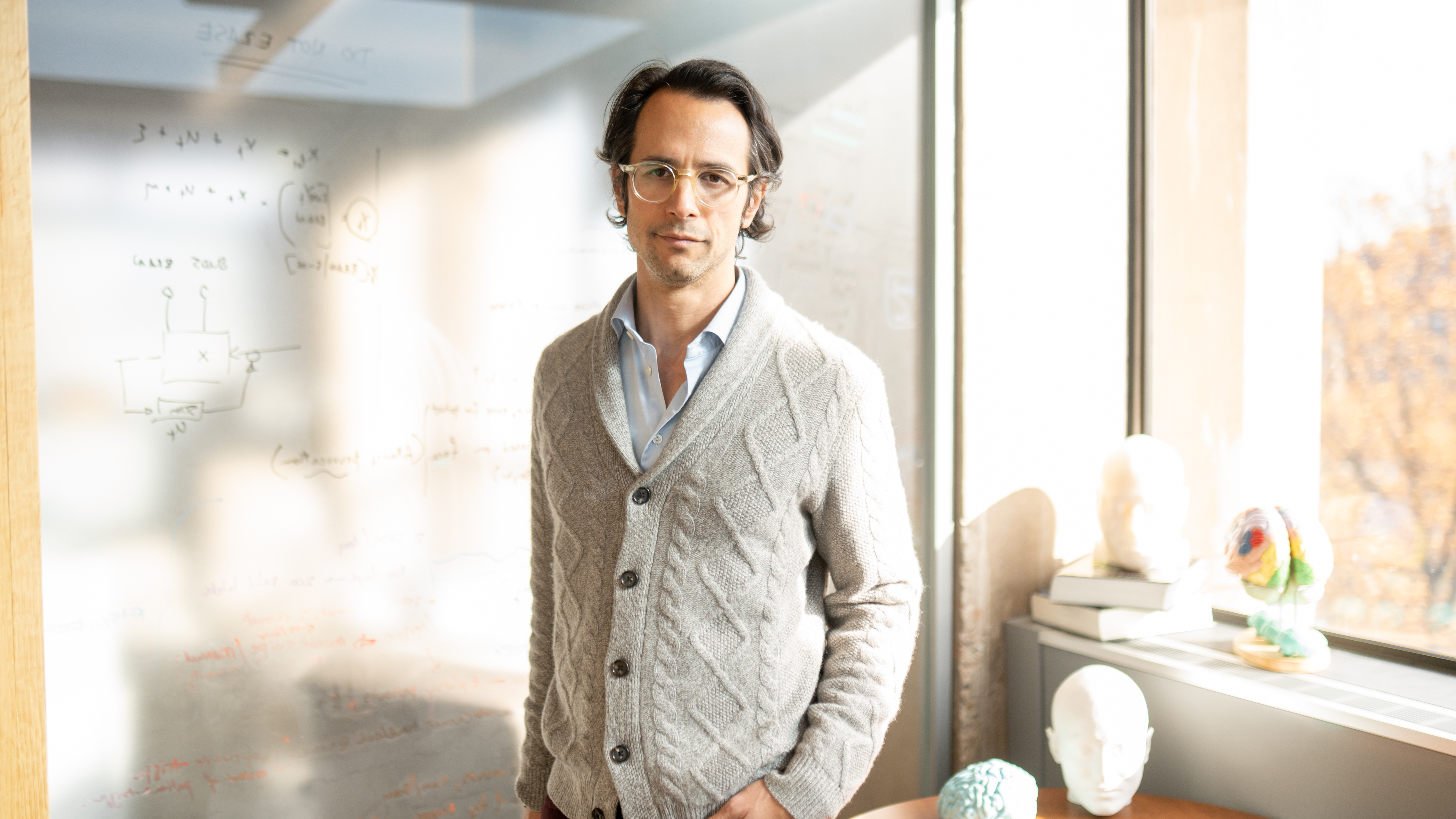 Man wearing glasses stands in front of white board with writing on it, and models of brains and skulls on a stack of books behind him
