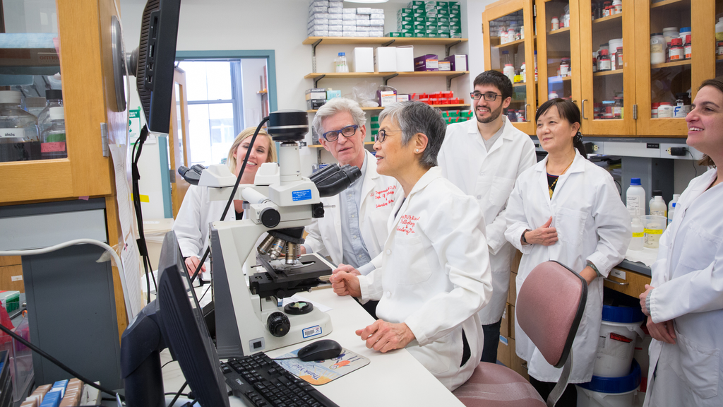 In a lab, John Q. Trojanowski looks toward Virginia Lee, as Lee and a surrounding group of lab members look at a screen in front of her.