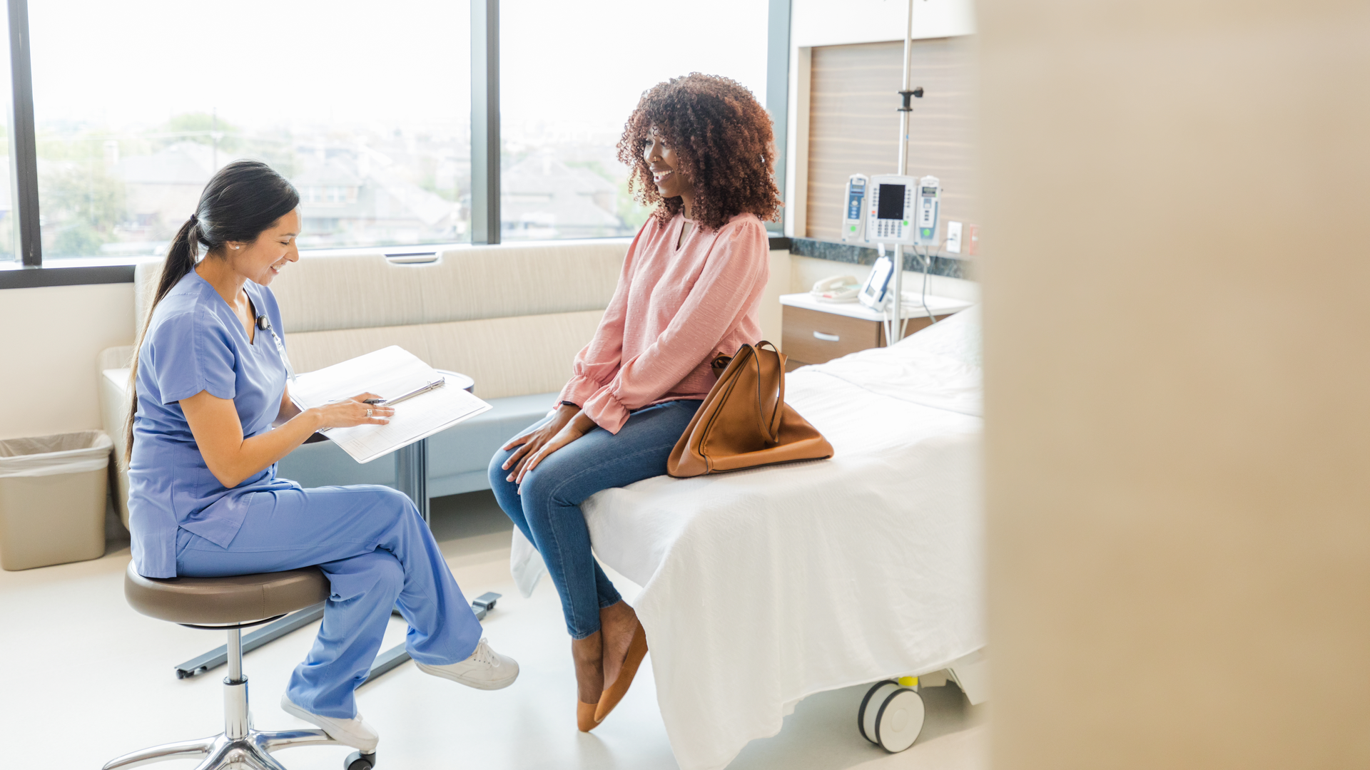 A doctor in a hospital room speaking to a patient sitting at the end of a bed, smiling.