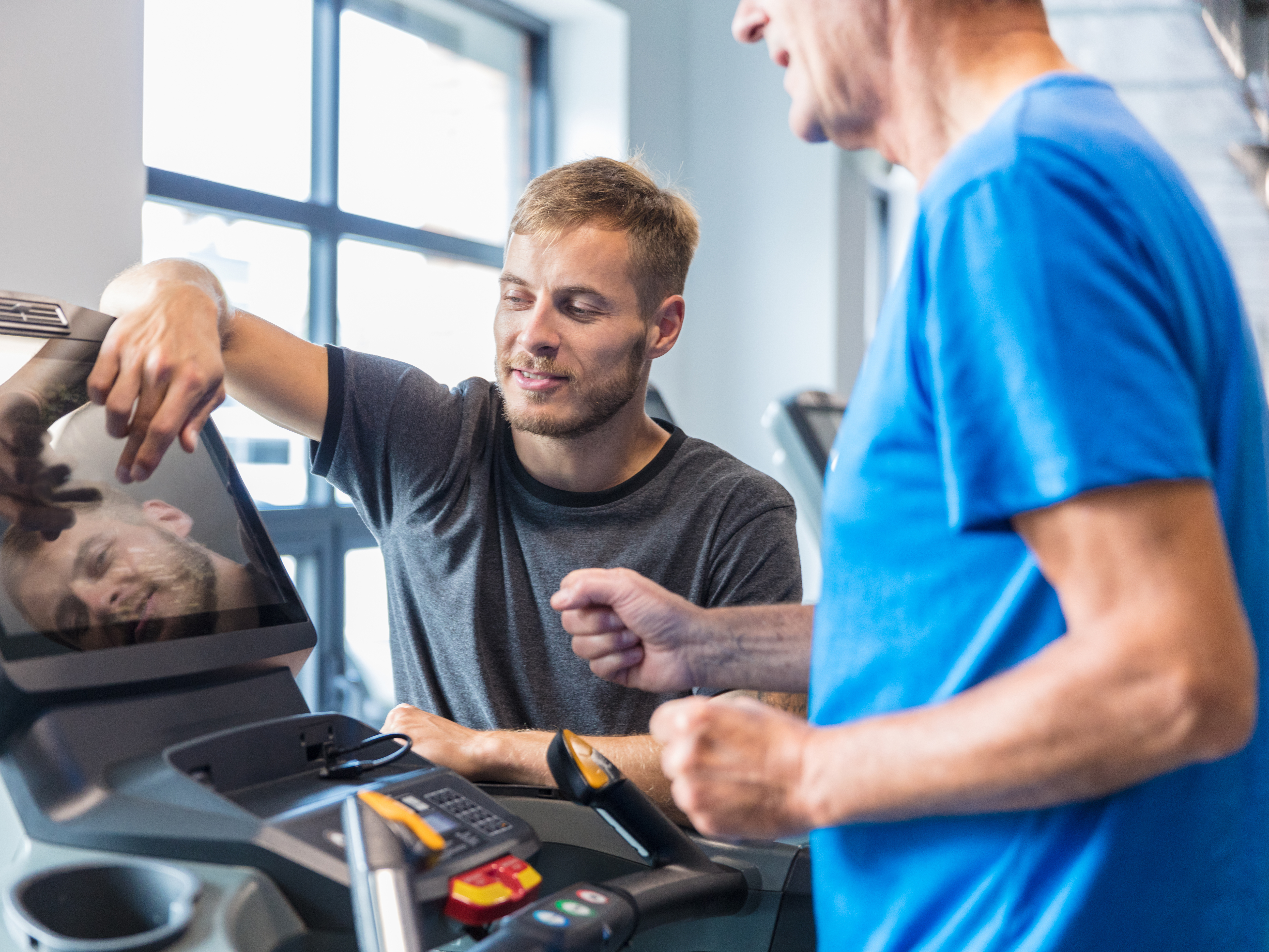 Trainer supervising older man on treadmill and explaining adjustments at rehab gym