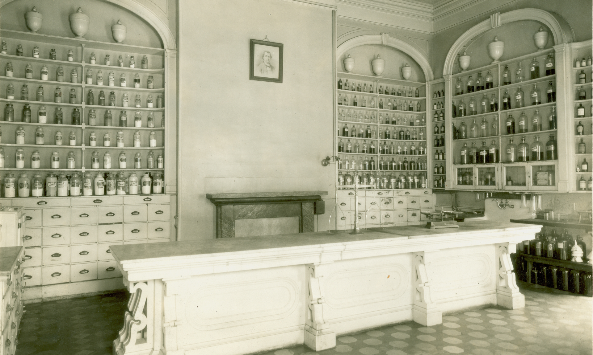 A black and white photo of the old Pennsylvania Hospital Apothecary shows a large marble or stone desk with white-painted drawers behind on the wall, with arched alcoves above the drawers lined with shelves of jars of liquids and bottles of powders, and a fireplace mantel in the center of the wall