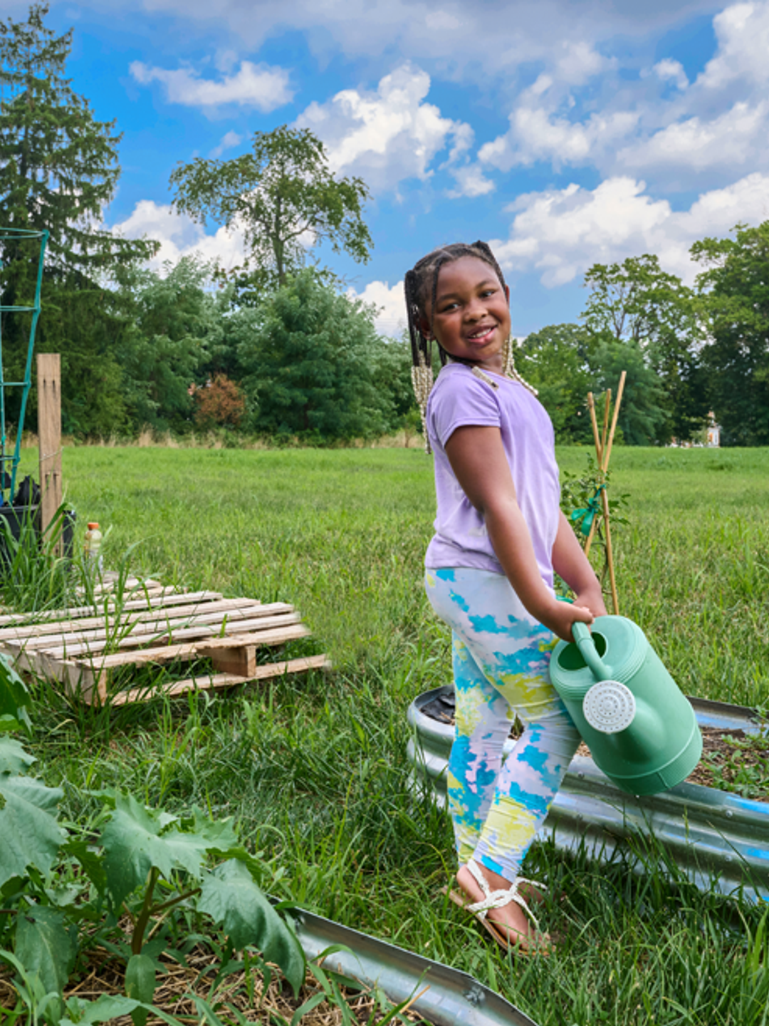 Young Black girl holding a watering can in front of urban garden