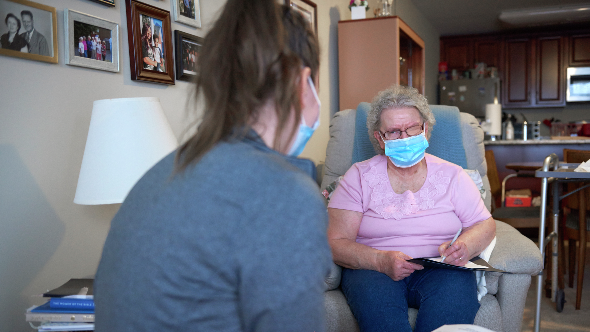 An older woman sits in an armchair, writing on a piece of paper, while looking at a nurse in scrubs facing her