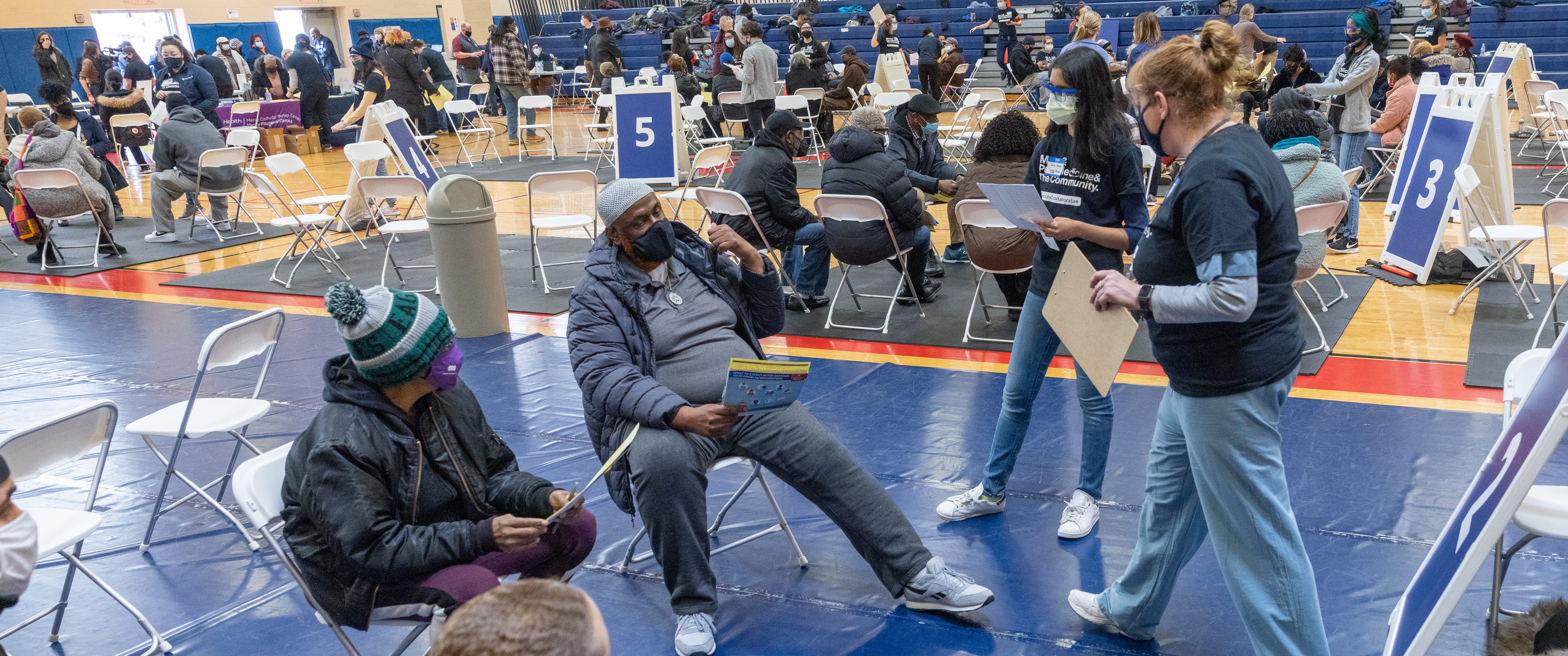 A school gym filled with people gathered in small groups of folding chairs, with standing people in navy T-shirts and scrub pants talking with them