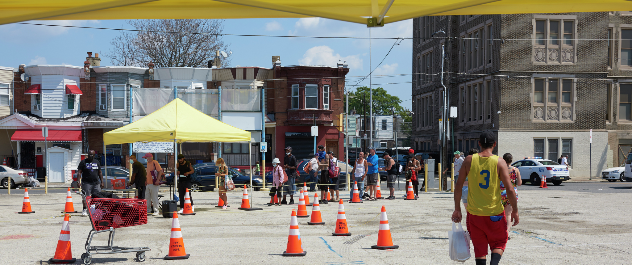 People gather in a line in an urban parking lot, as they approach a yellow pop-up tent