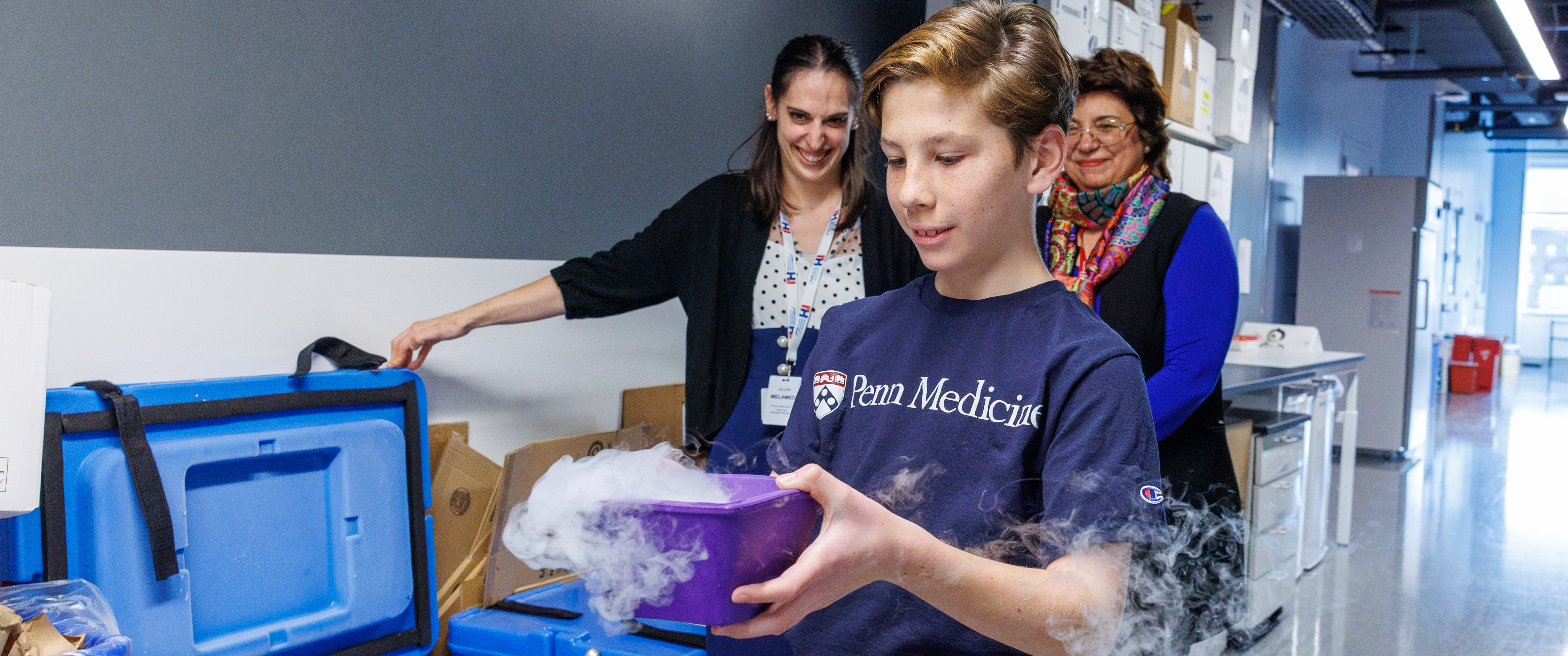 11-year-old Jax Bari carries a purple bin emitting vapor as if frozen, in a lab hallway, as Jilian Melamed and another person look on
