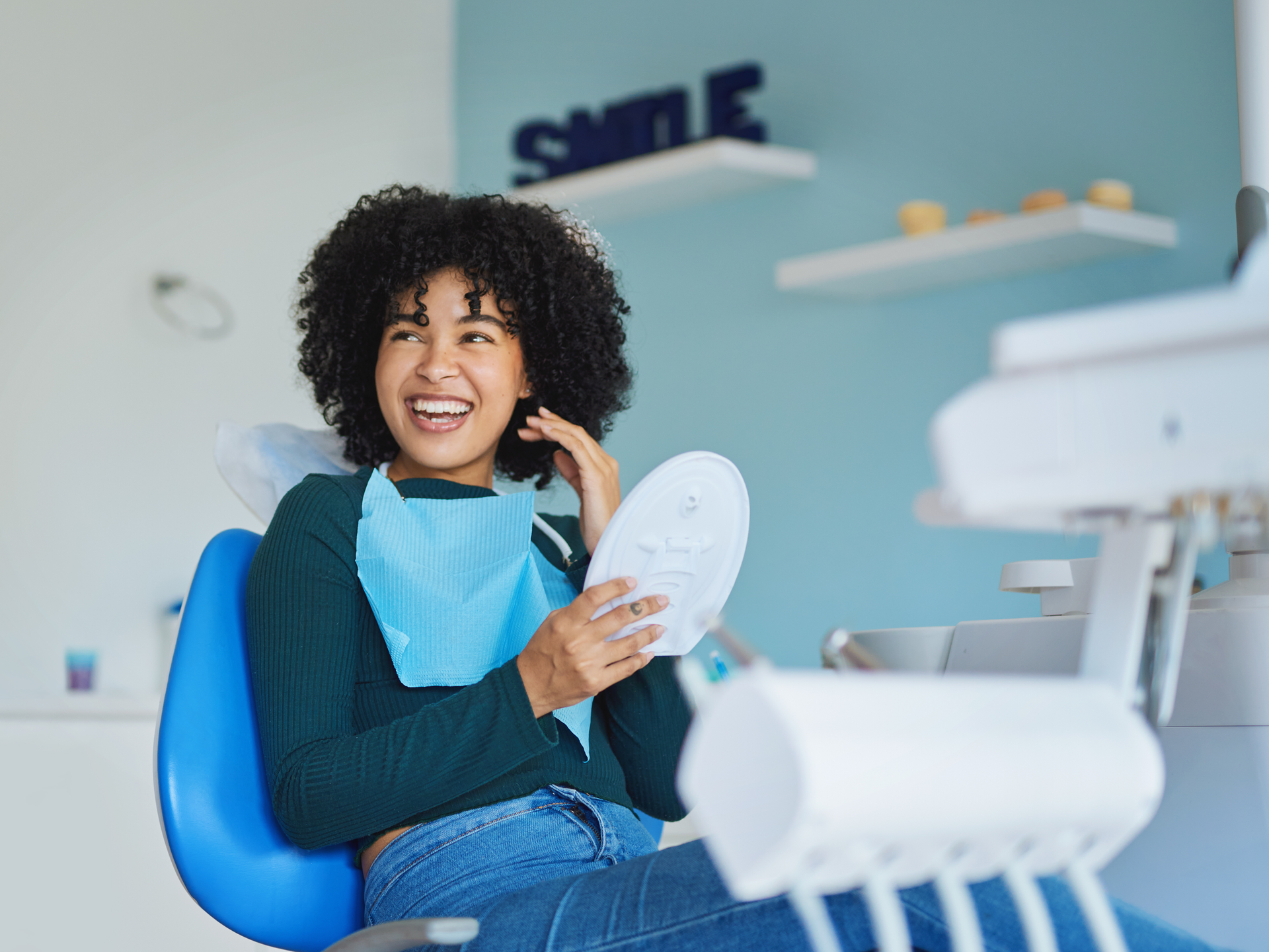 A dental patient smiling at someone just out of frame as she holds a mirror up to look at her teeth