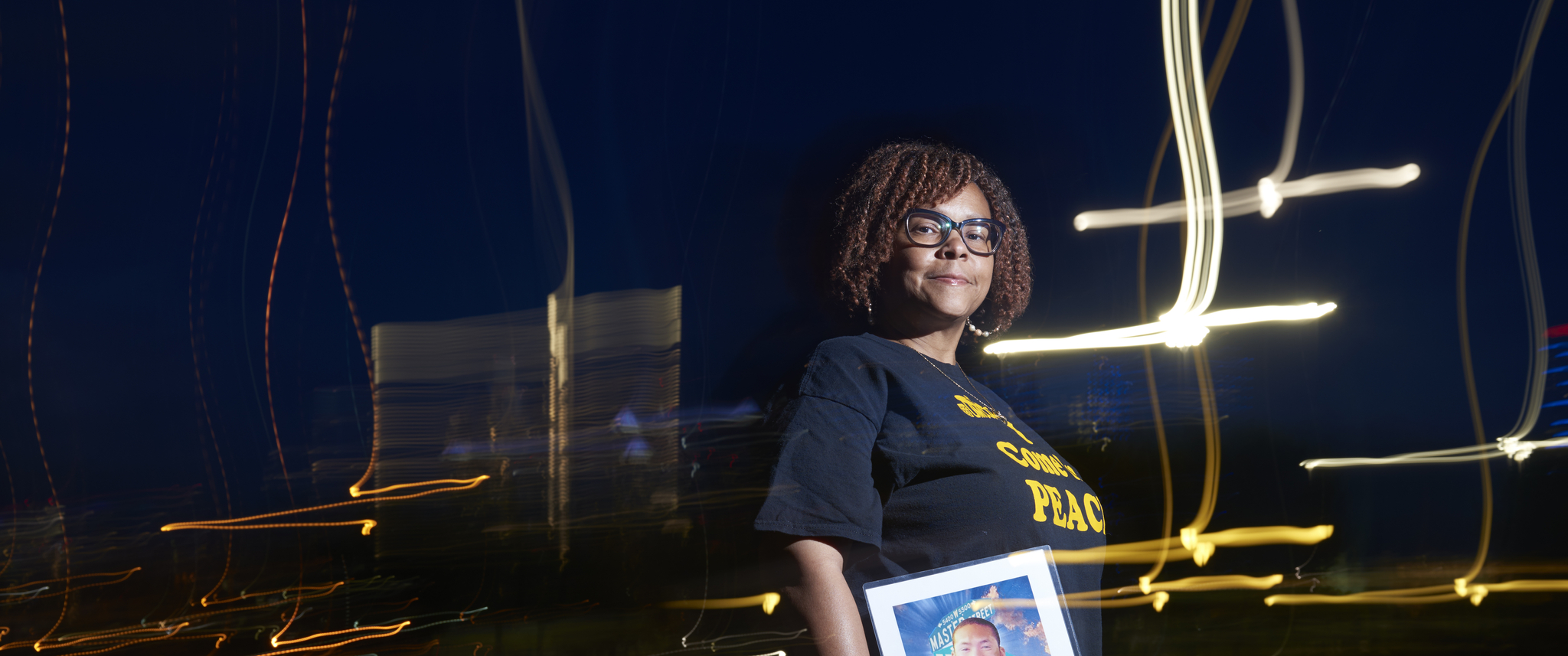 Cheryl Graham-Seay, at night with a blurred city skyline behind her, stands facing the viewer, holding a photo of her son