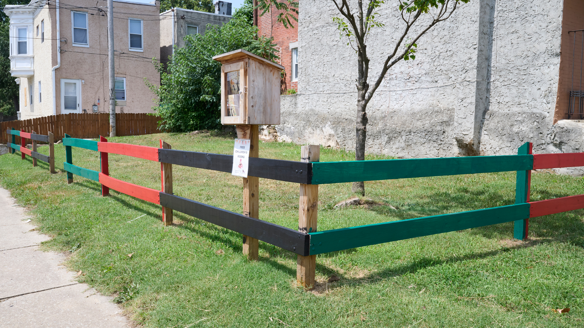 A grassy corner lot adjacent to rowhouses is surrounded by a painted wooden fence, with a little free library kiosk in front