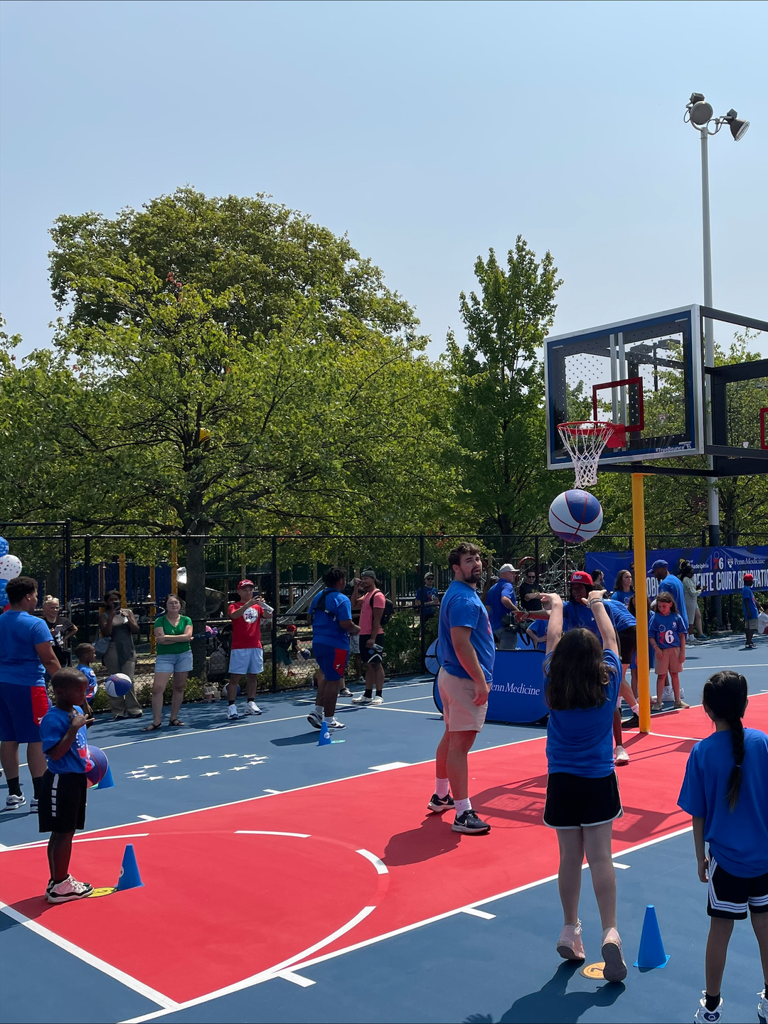 A girl shoots a basketball with families gathered, in Philadelphia Sixers t-shirts, on the new basketball court in Roberto Clemente Park