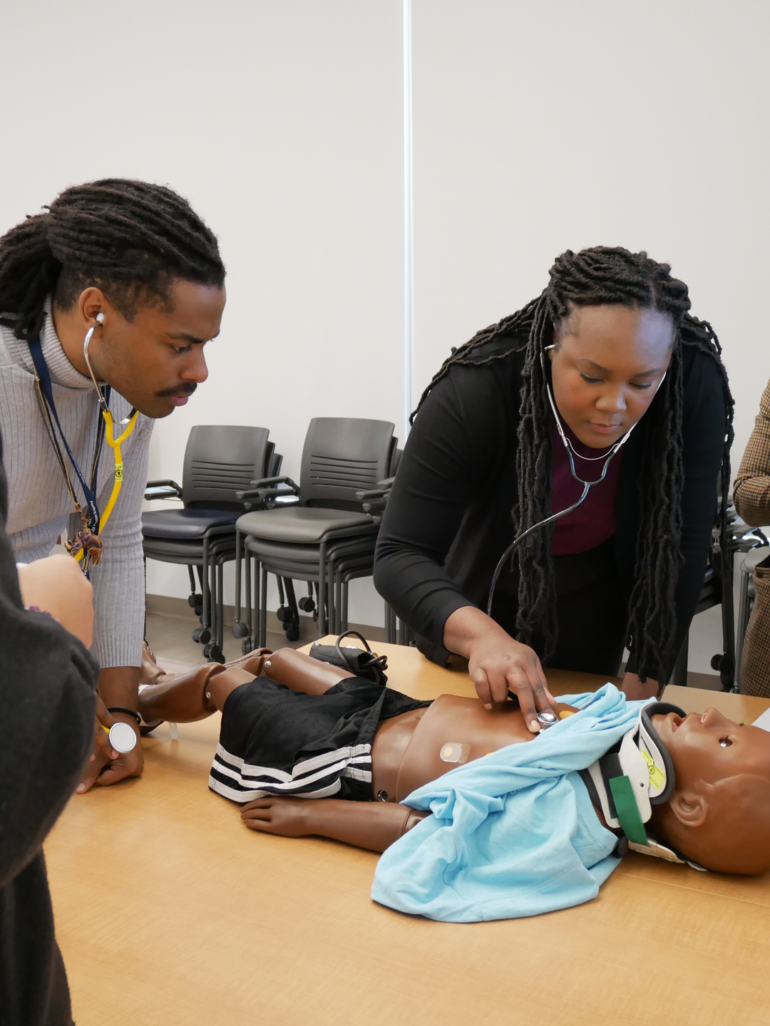 Claudia Gambrah-Lyles, MD, wearing a tweed blazer and holding a tablet, watches as three Black medical students monitor a child-sized manikin representing a trauma patient.