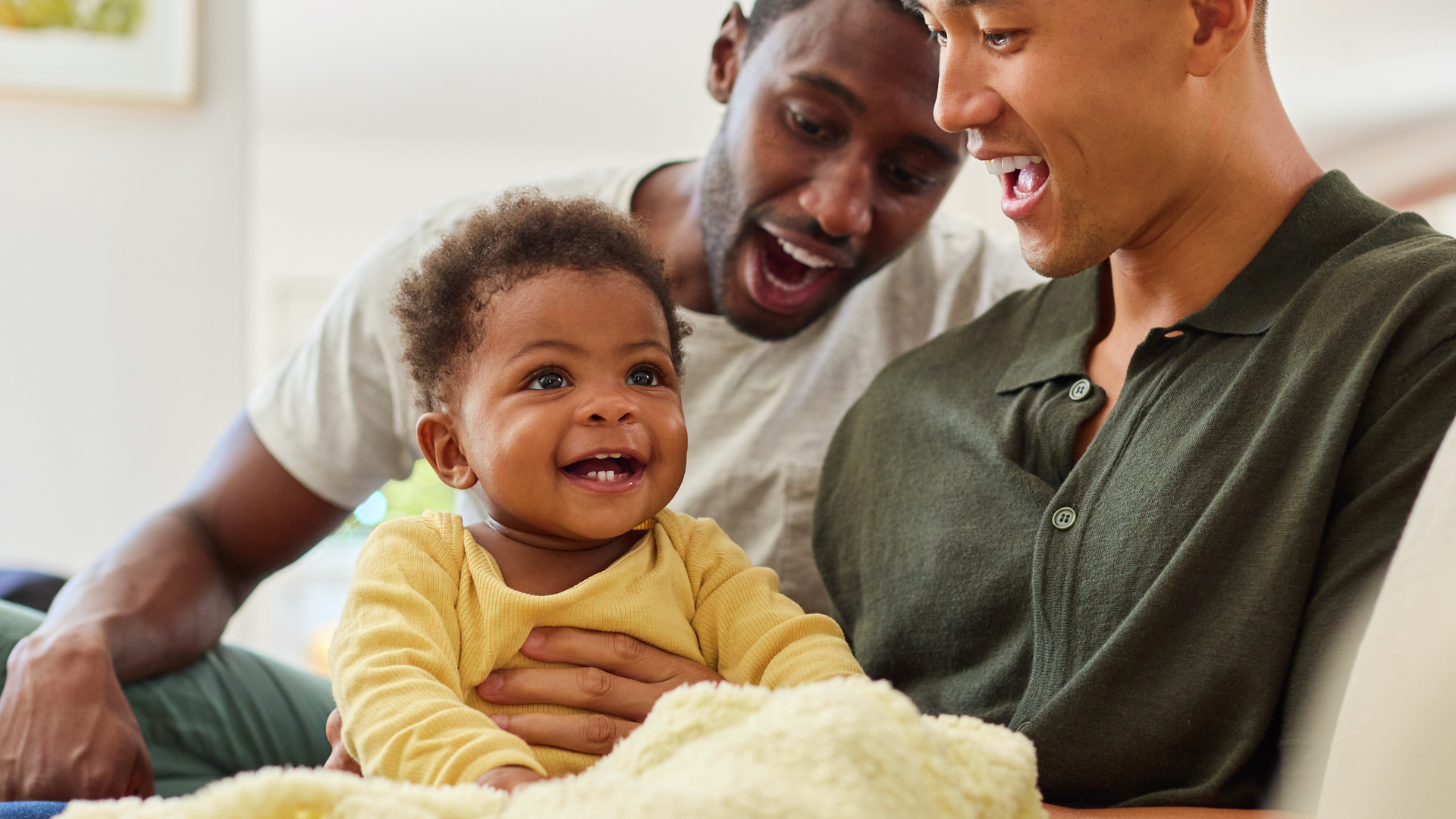 Same sex male couple or friends playing with baby sitting on sofa at home together