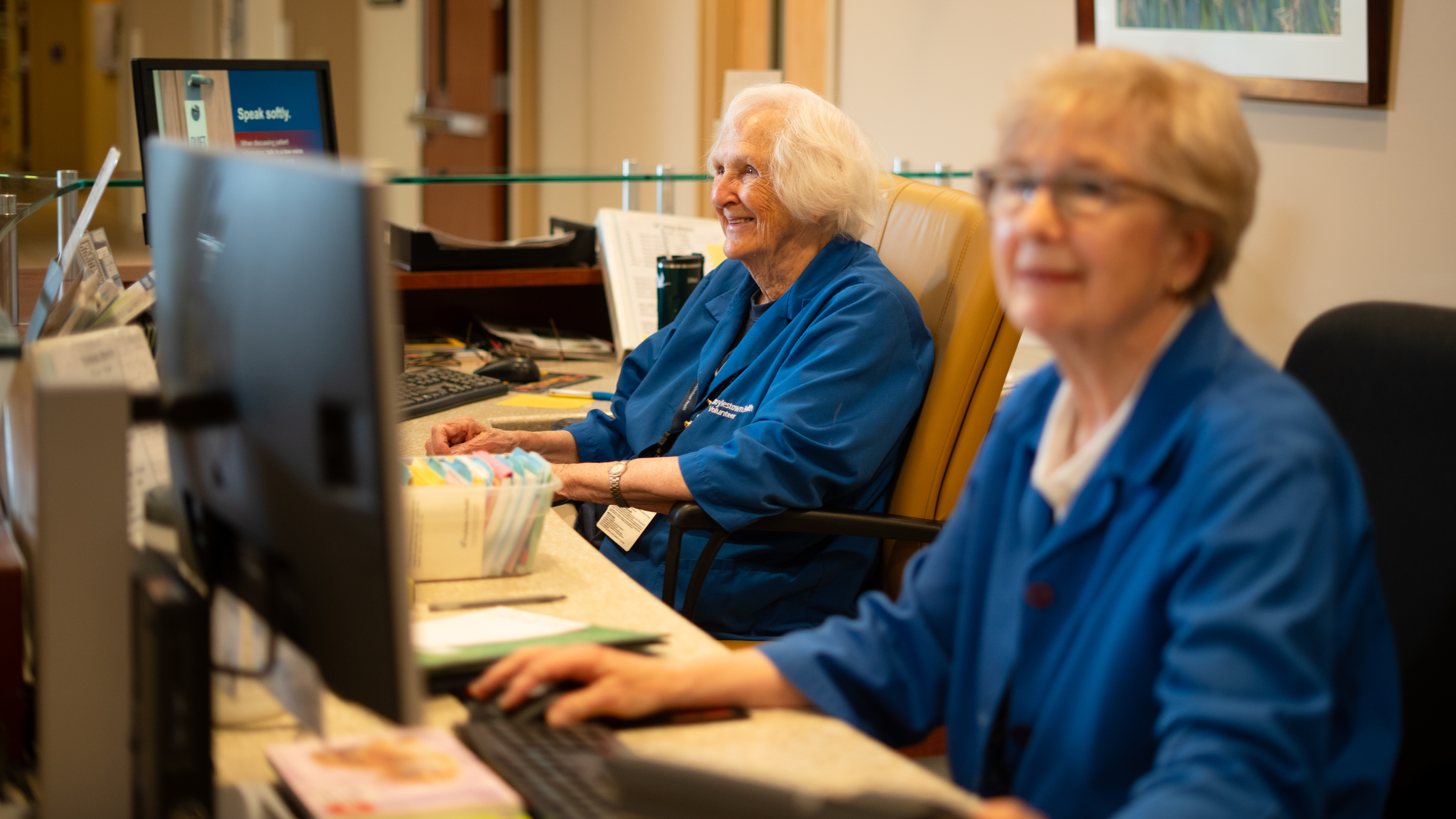 Carol Duffy and Carol Ermolde, seen sitting at their desk and smiling, working as information desk volunteers 