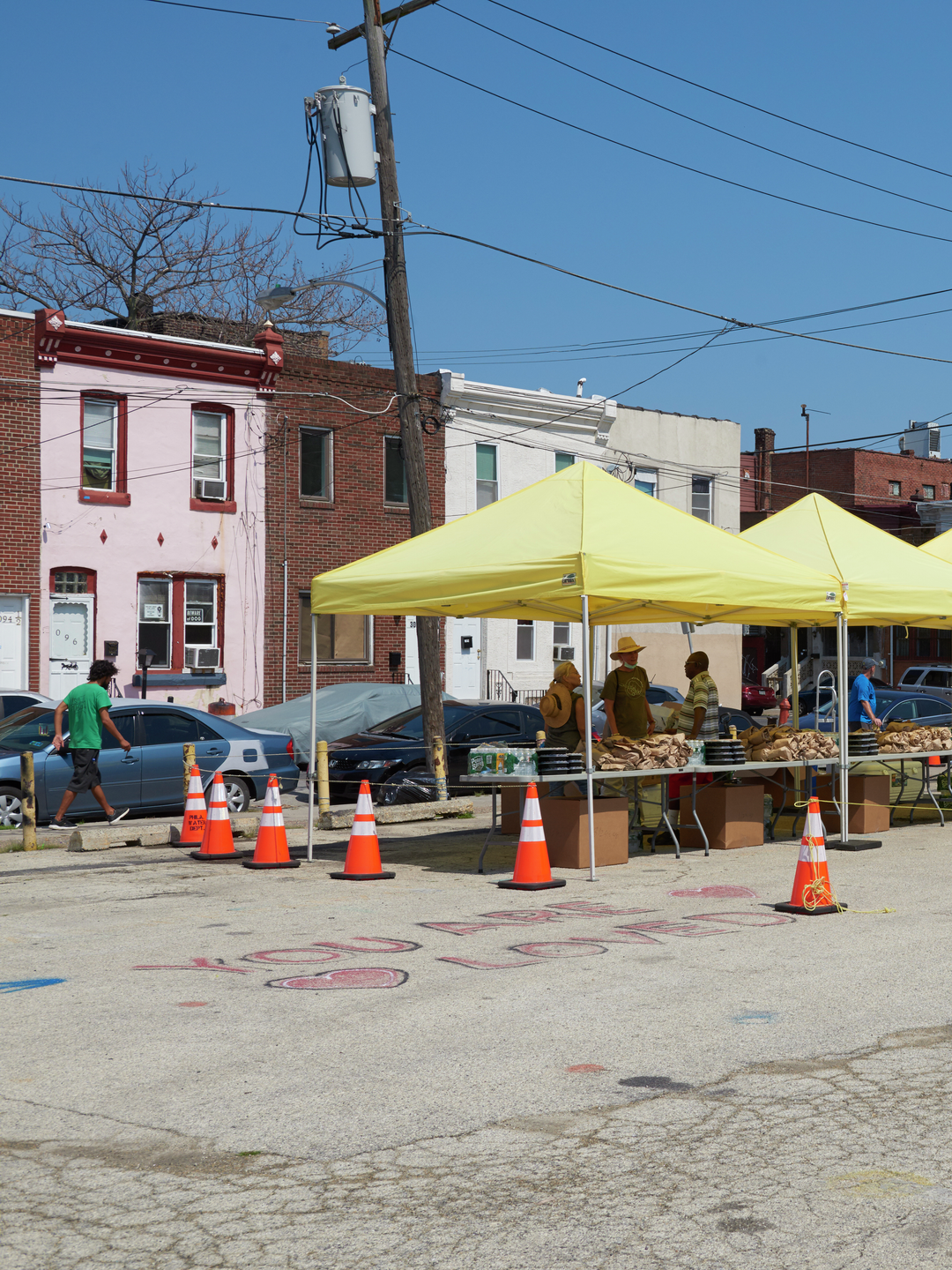 Under a yellow tent on a sunny summer day, a folding table is covered with plastic containers and paper bags of free food