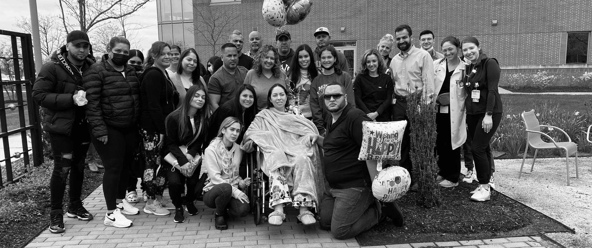 A group of about 30 people, some hospital staff in uniform, some holding birthday balloons, with a young woman seated in a wheelchair at the center