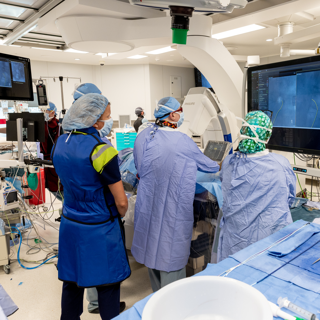 Two people wearing gowns, masks, and surgical bonnets manipulate machinery in an OR, facing screens