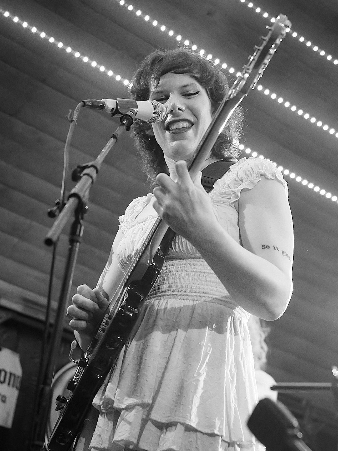 Black and white photo of Kimi Klauser holding an electric guitar and standing in front of a microphone, smiling, in a tiered white dress